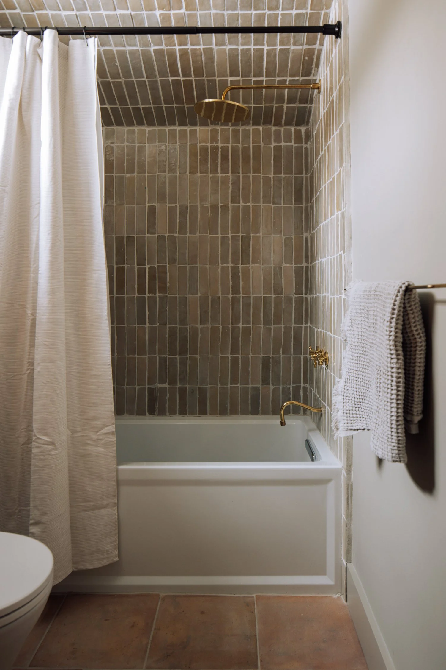 Bathroom shower with beige brick tiles, a white bathtub, a beige shower curtain, gold-colored shower fixtures, and a beige towel hanging on a towel rack.