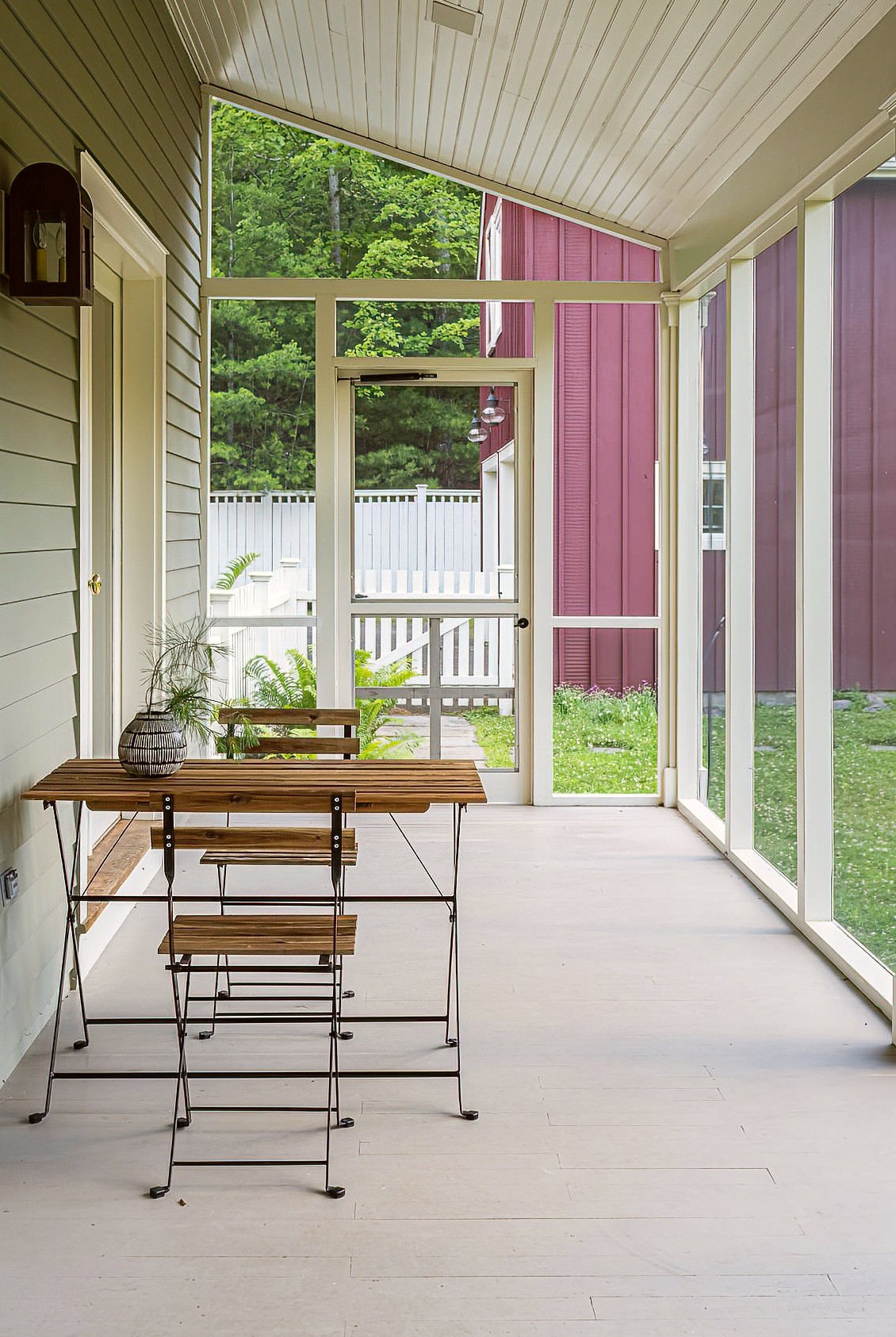 Enclosed porch with a small wooden table and chairs, potted plants, and large windows showing a yard with a white fence and trees.