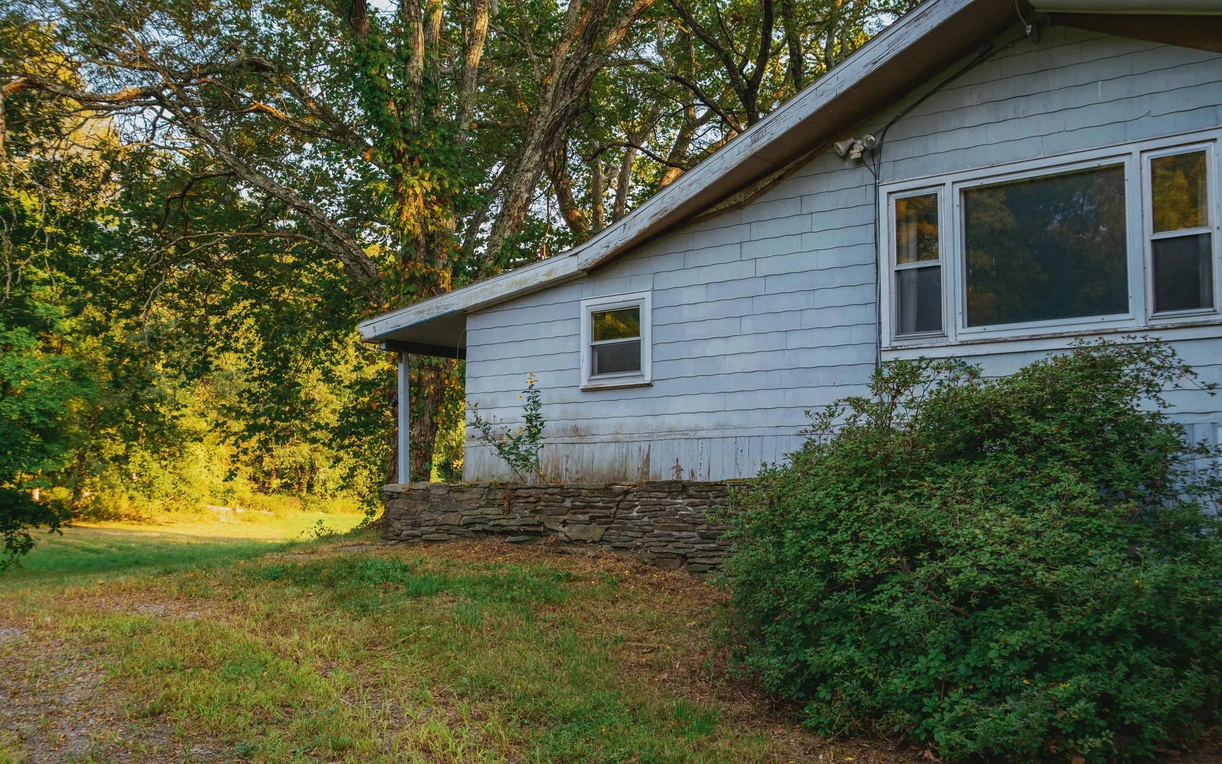 A white house with a sloped roof surrounded by trees and greenery, with a stone foundation and small windows.