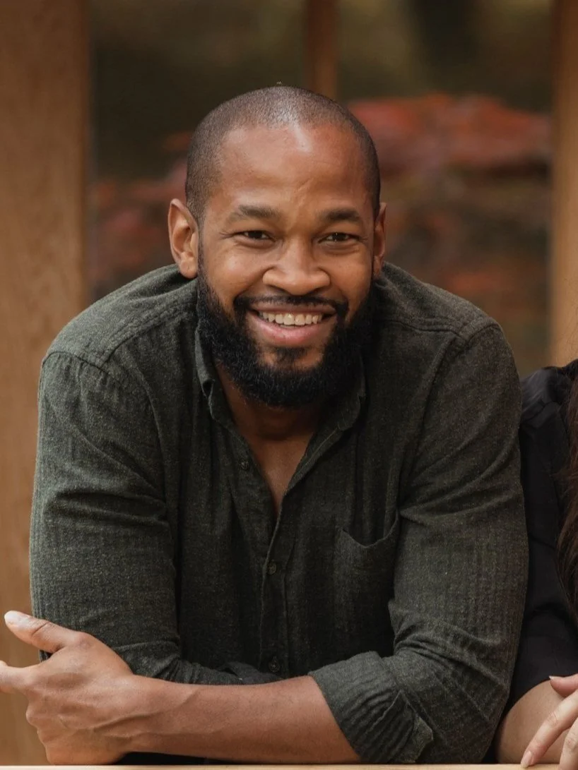 A smiling man with a beard and short hair, dressed in a dark green shirt, resting his arms on a surface in an outdoor setting.