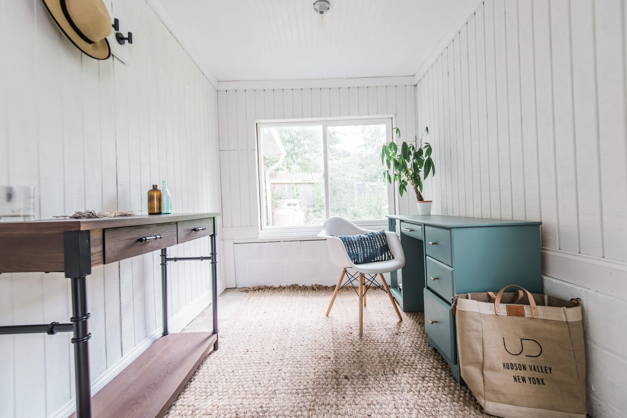A small room with white paneled walls and a window, decorated with a blue desk, a white chair with a blue cushion, a green potted plant, a beige storage bag, a wooden side table with bottles, and a straw hat hanging on the wall.