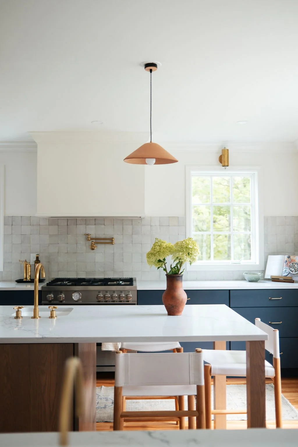 Kitchen with white walls, a window, a hanging terracotta-colored pendant light, a marble island with a vase of white hydrangeas, blue lower cabinets, gold fixtures, and a wooden dining chair.
