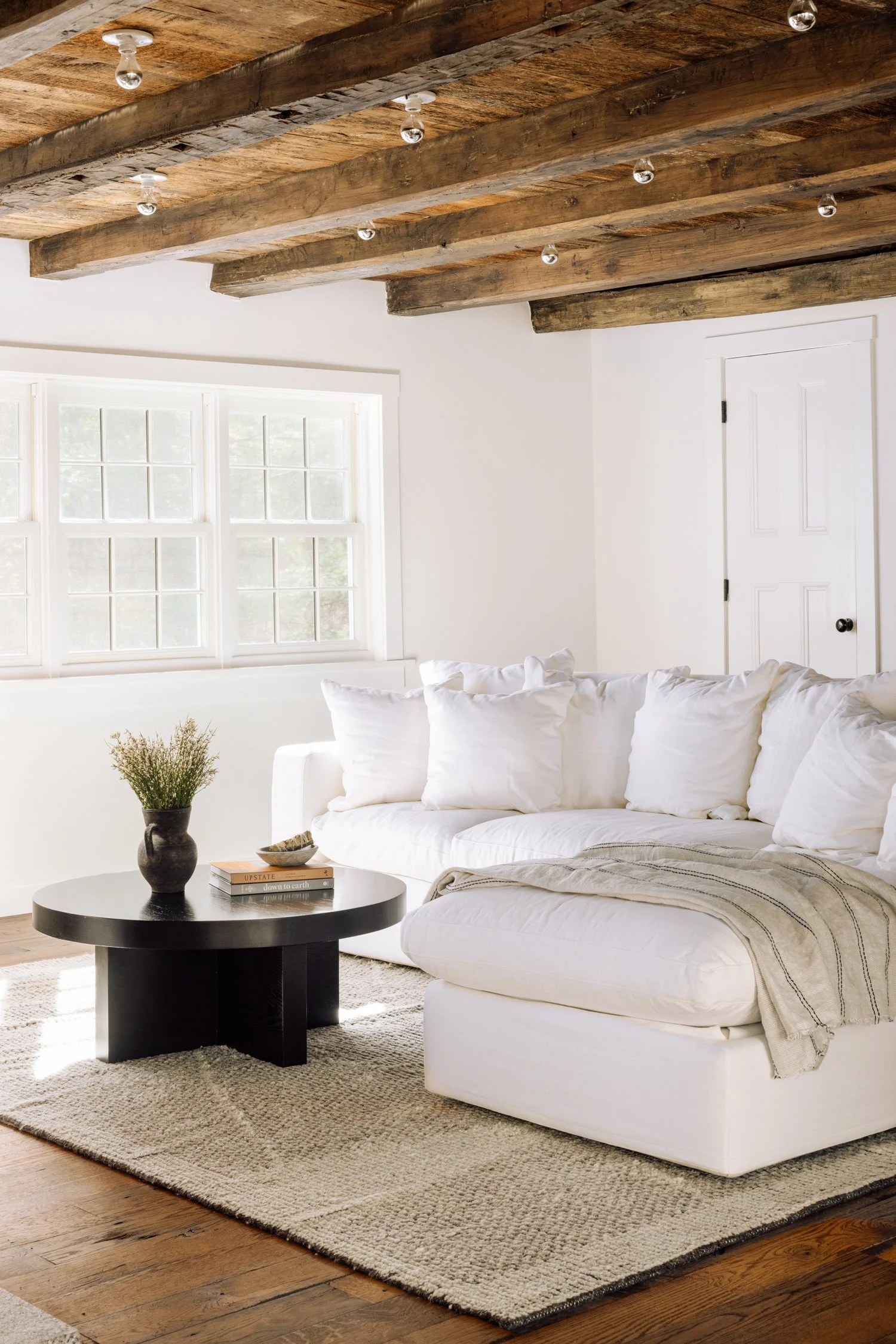 A cozy living room with a white sectional sofa, black coffee table with books and a vase, a textured rug, large windows, and exposed wooden ceiling beams.