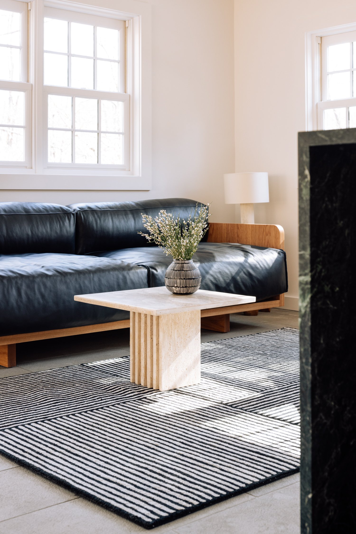 Living room with black leather sofa, wooden side table with a striped vase and flowers, white walls, and a black-and-white striped rug near a window.