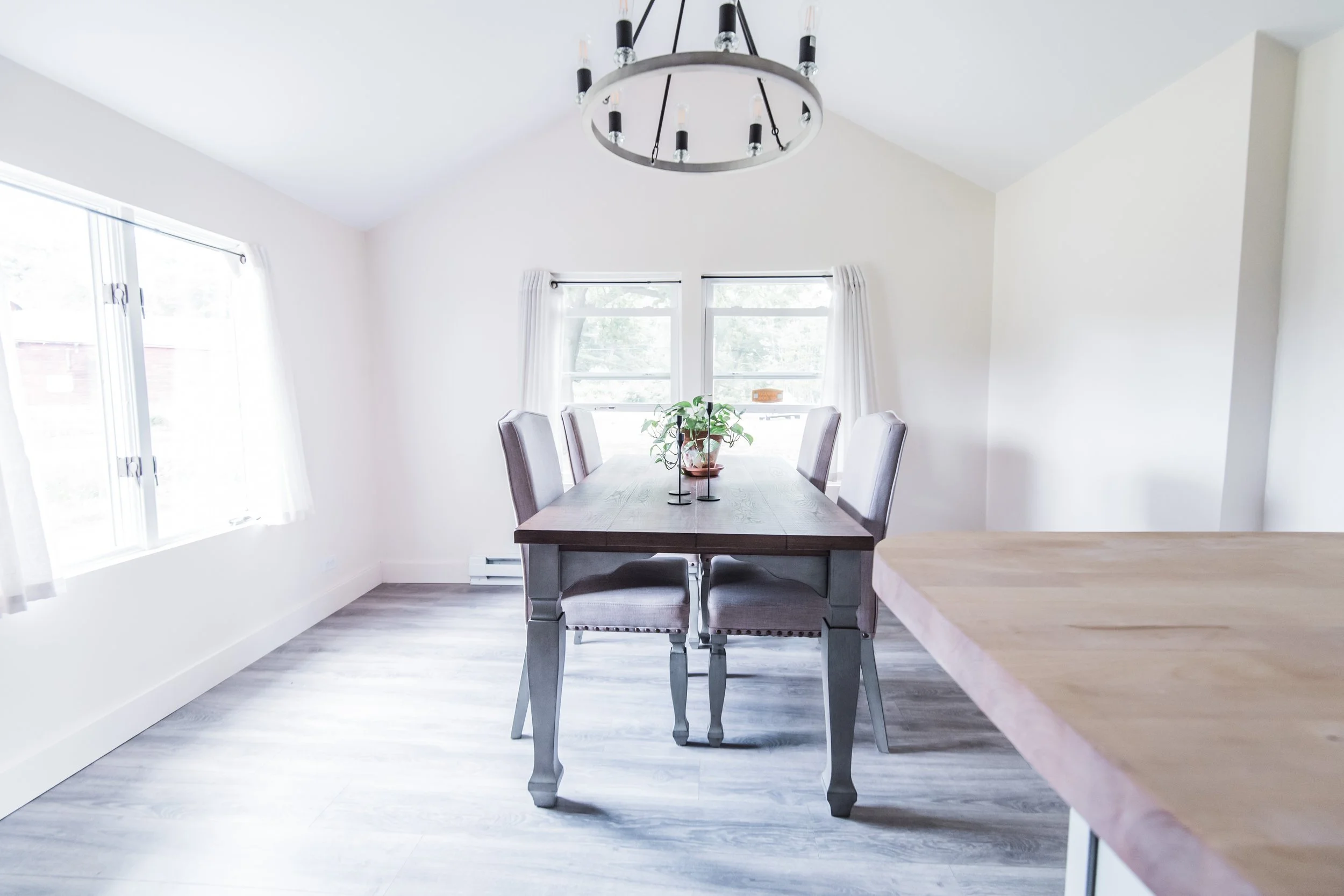 Empty dining room with a wooden table, six chairs, a plant centerpiece, large windows with white curtains, and a modern circular chandelier.
