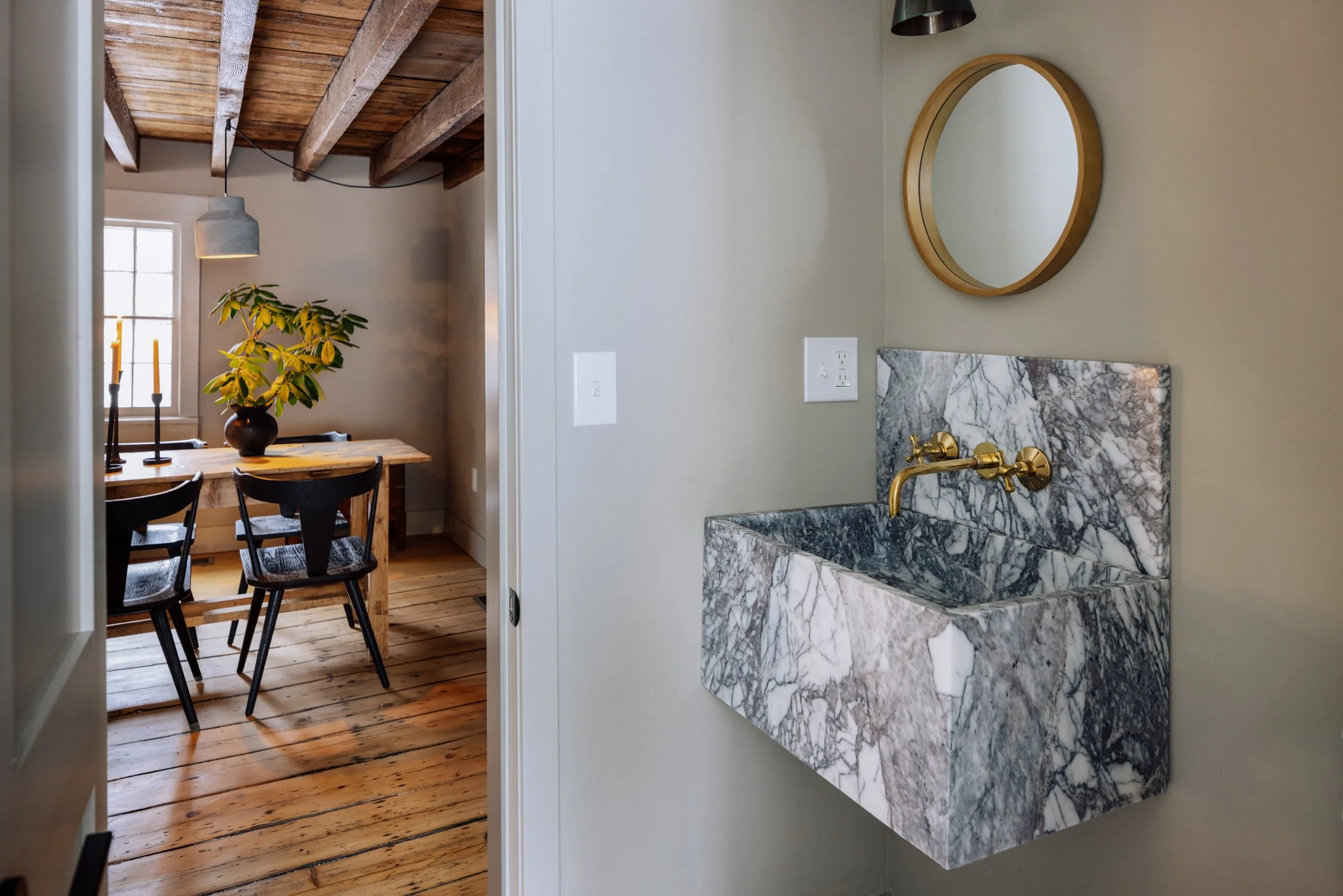 Bathroom with a marble sink, brass faucet, and round mirror on a beige wall. Adjacent dining room with wooden table, black chairs, a window, and exposed wooden beams on the ceiling.