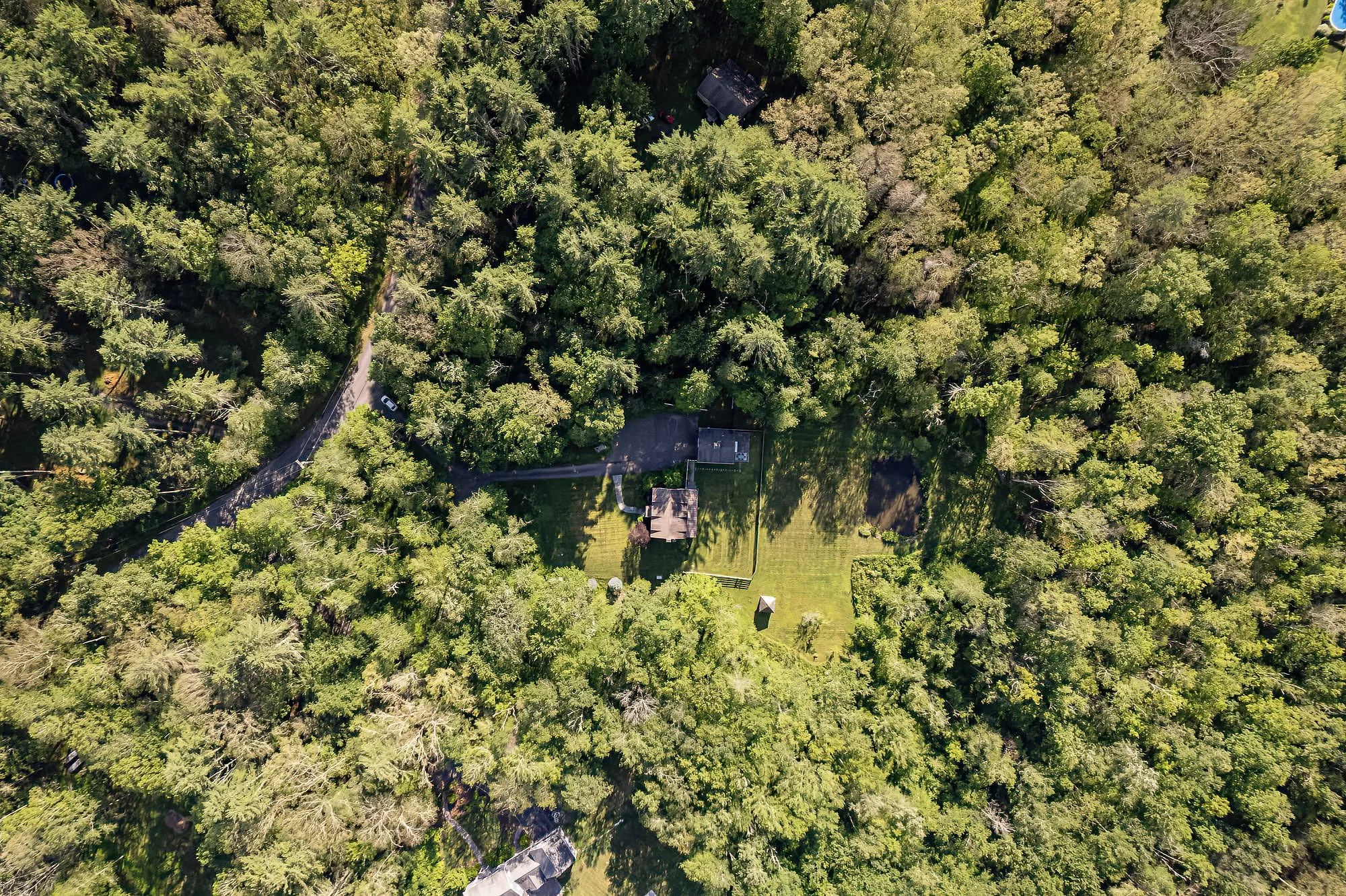 Aerial view of a house surrounded by dense trees with a lawn and driveway.