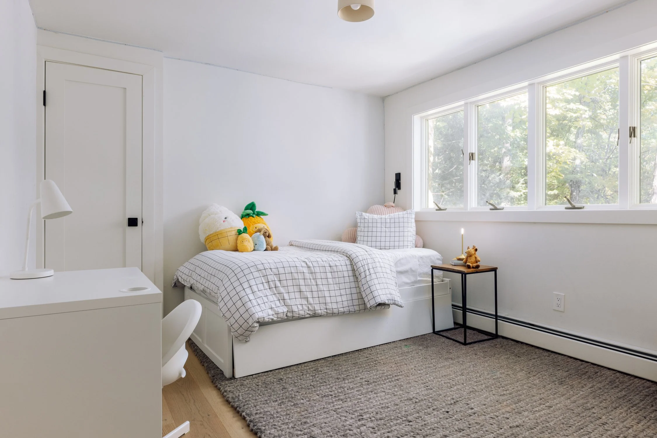 Minimalist bedroom with white walls, a bed with checkered bedding, plush toys, a small black side table with a teddy bear, and large windows showing trees outside.