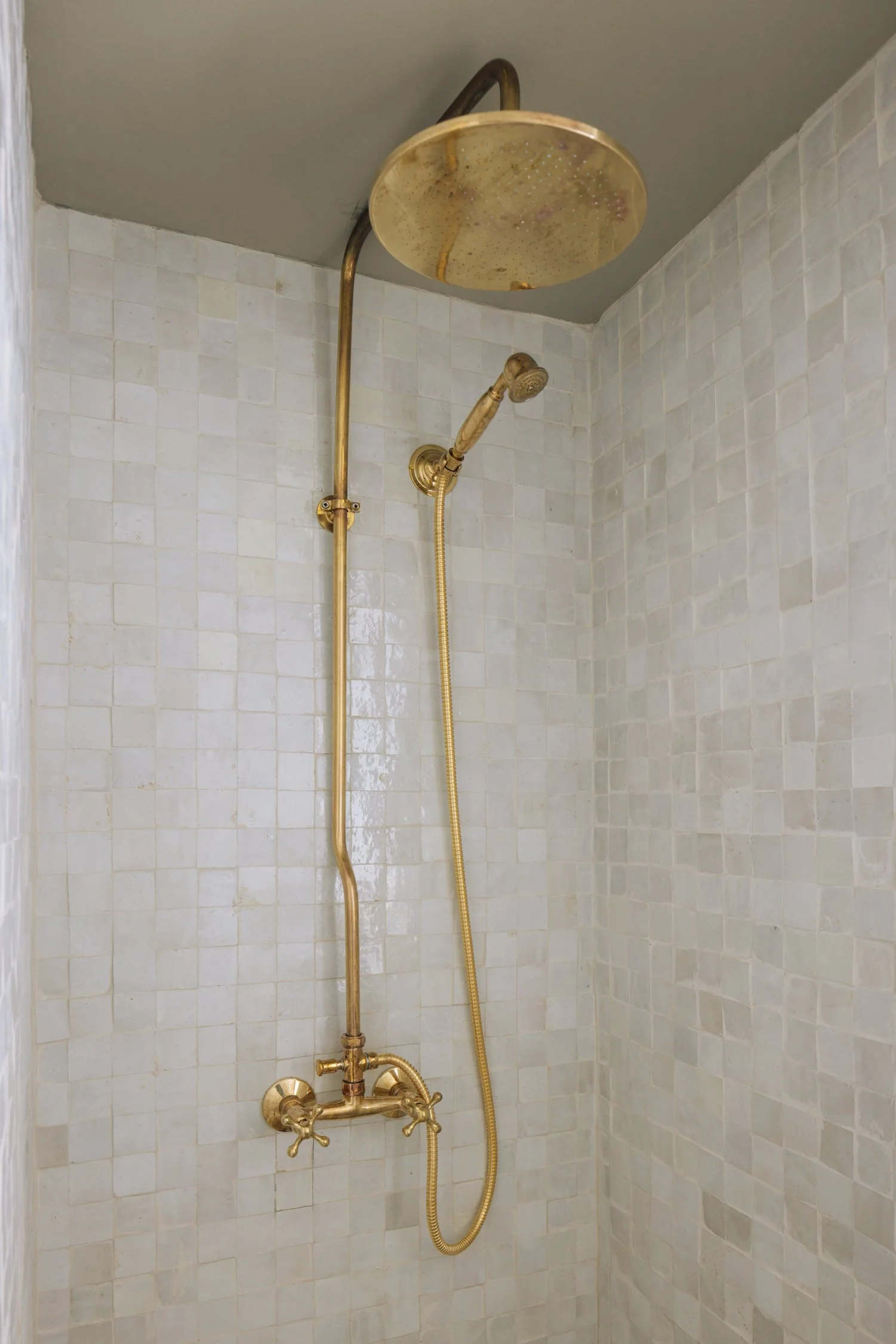A shower with brass fixtures, a large overhead showerhead, and a smaller handheld showerhead, against tiled walls in a bathroom.
