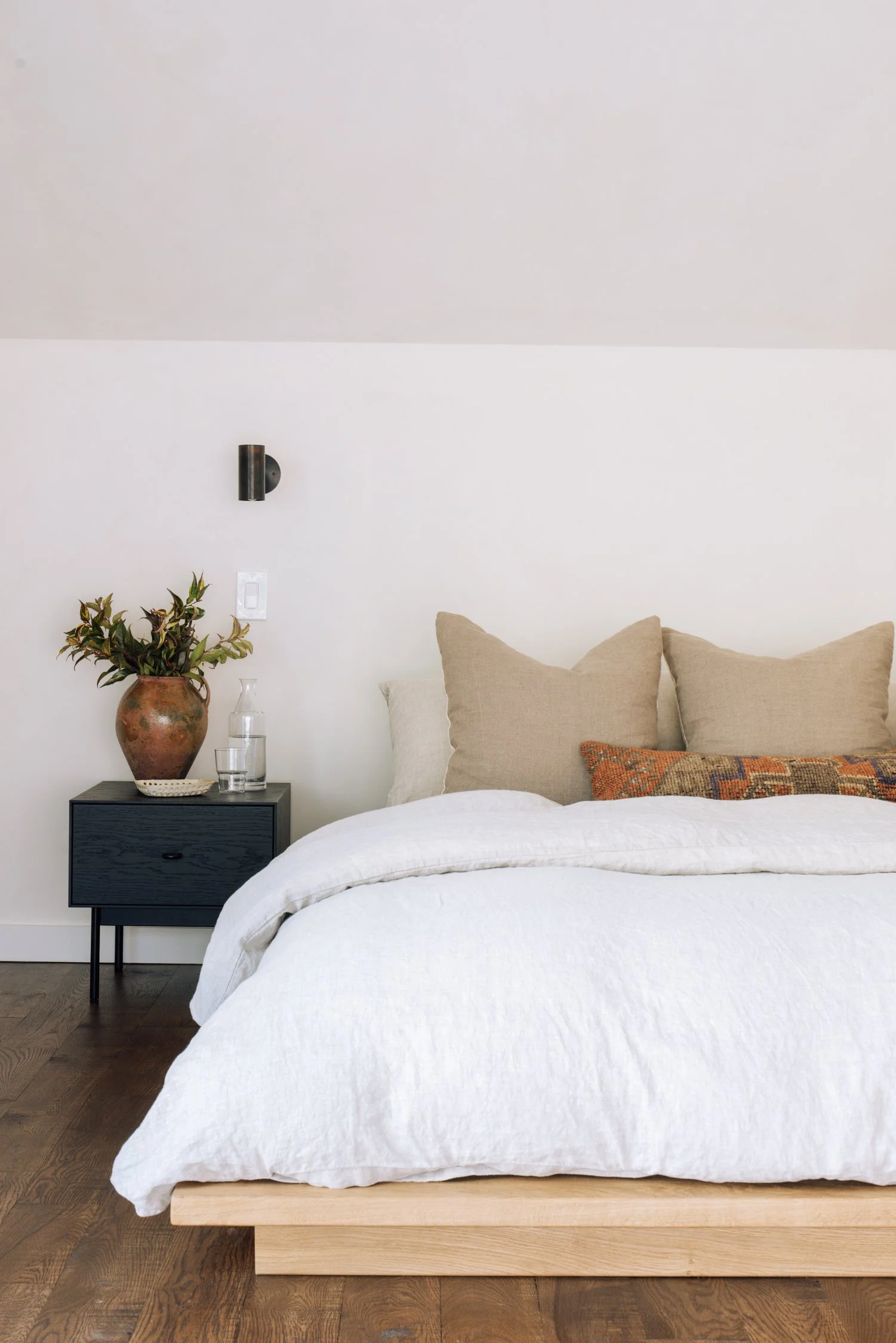 A bedroom with a bed that has beige and orange pillows, a white bedspread, a black nightstand with a clay vase, a glass, and a small decorative object, and a wall-mounted black sconce light on a white wall.