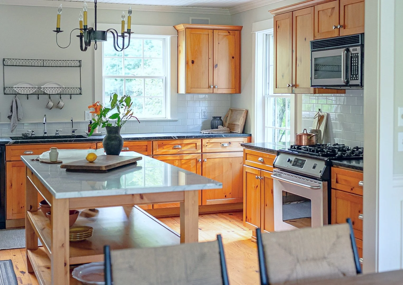 Kitchen with wooden cabinets, black countertops, white subway tile backsplash, stainless steel appliances, a kitchen island with a wooden shelf, vase with flowers on the island, and two windows providing natural light.