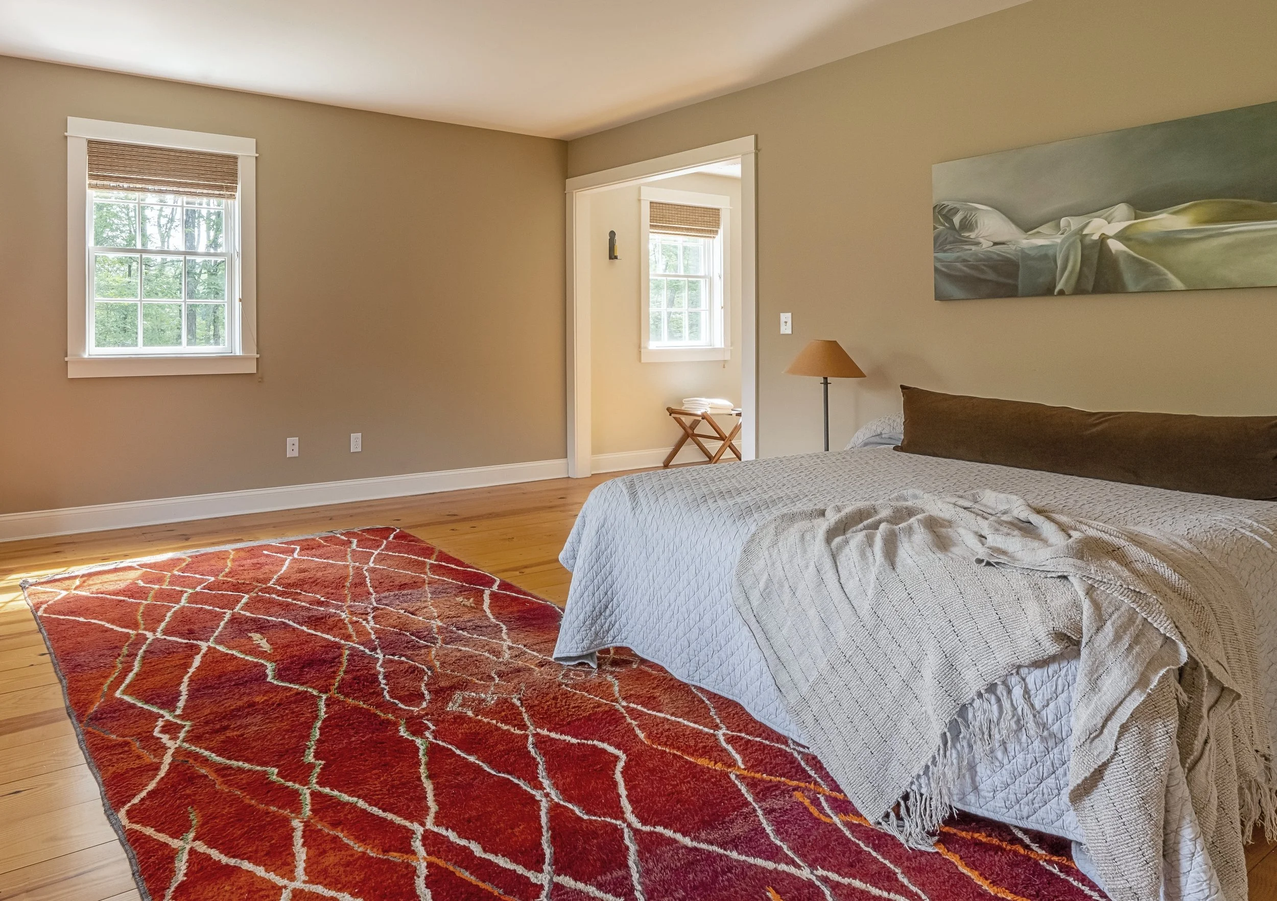 A bedroom with a large bed, beige walls, wooden floor, a colorful red and orange area rug, and two windows with blinds. There is a small bedside lamp, a framed artwork of a bed, and a small stool with towels in a corner.