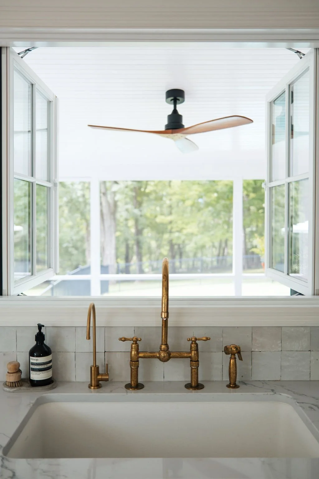 Kitchen sink with brass fixtures, a soap dispenser, and a window with a view of green trees outside. A ceiling fan with wooden blades is visible through the window.
