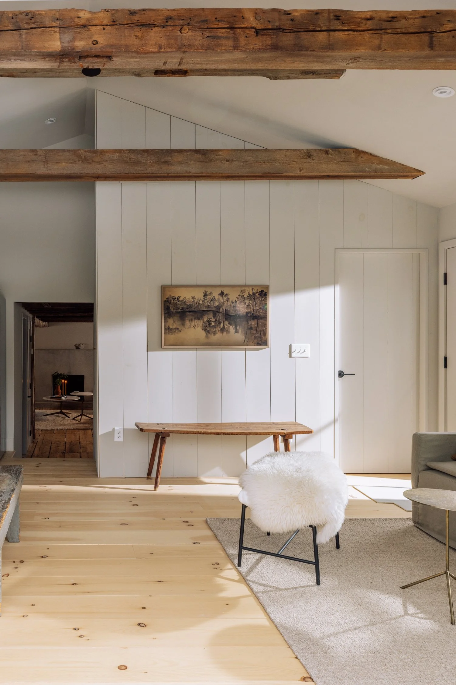 Living room with wooden beams on the ceiling, white paneled wall, a small wooden bench, a sheepskin-covered chair, a gray sofa, a light-colored area rug, and a framed landscape painting.