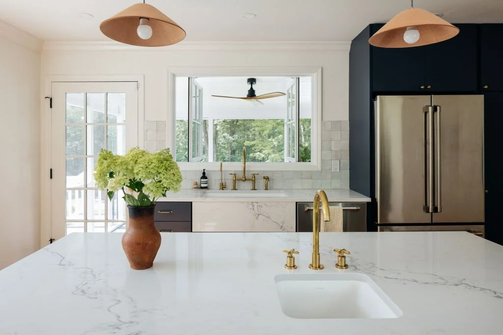 Modern kitchen with white marble island, gold fixtures, dark blue cabinets, stainless steel refrigerator, potted flowers, and open window showing trees outside.