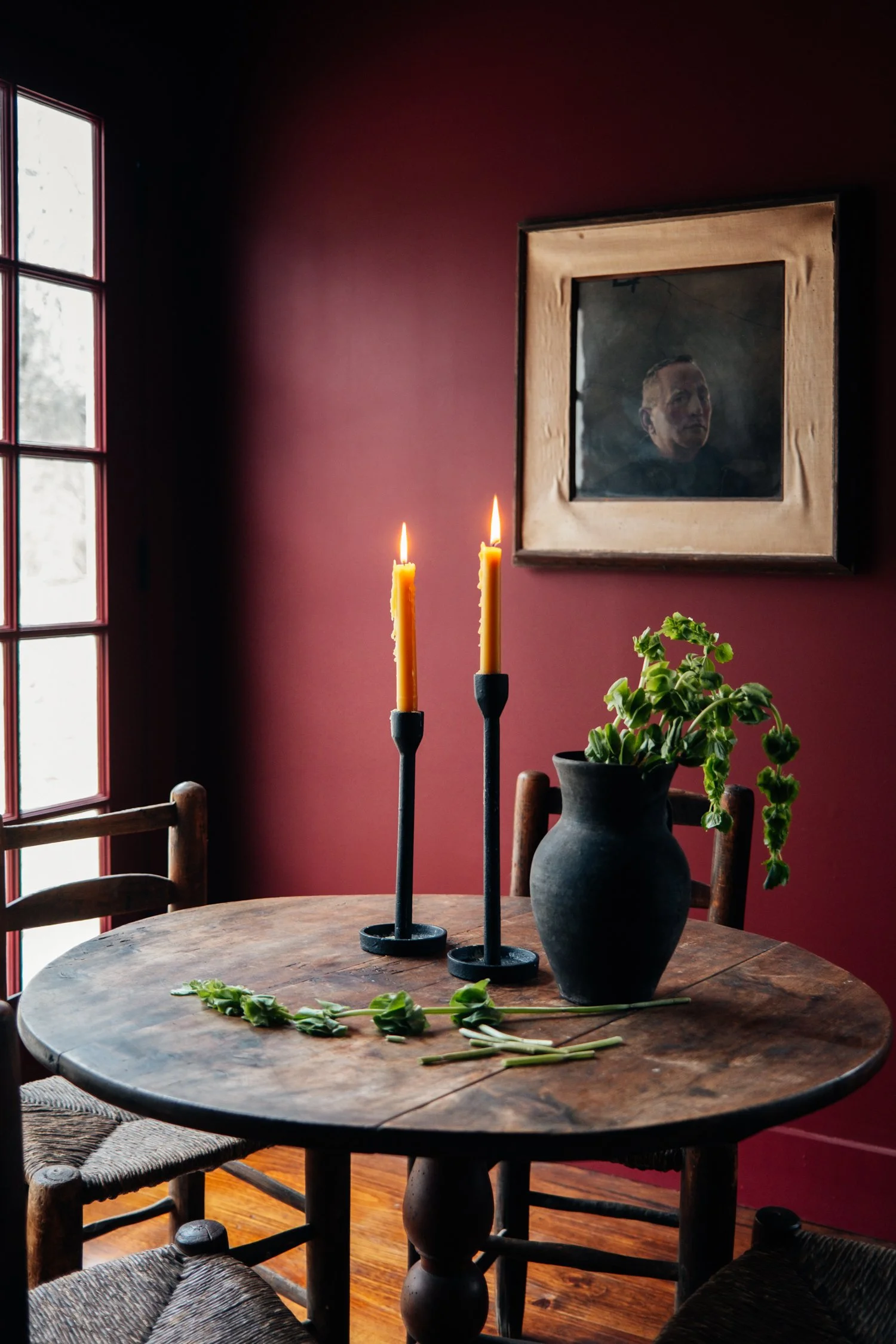 A round wooden table with two black candle holders holding lit yellow candles, a black vase with green foliage, and a few loose green leaves on top, in a room with red walls and a window with snow outside, with a picture of a man hanging on the wall.