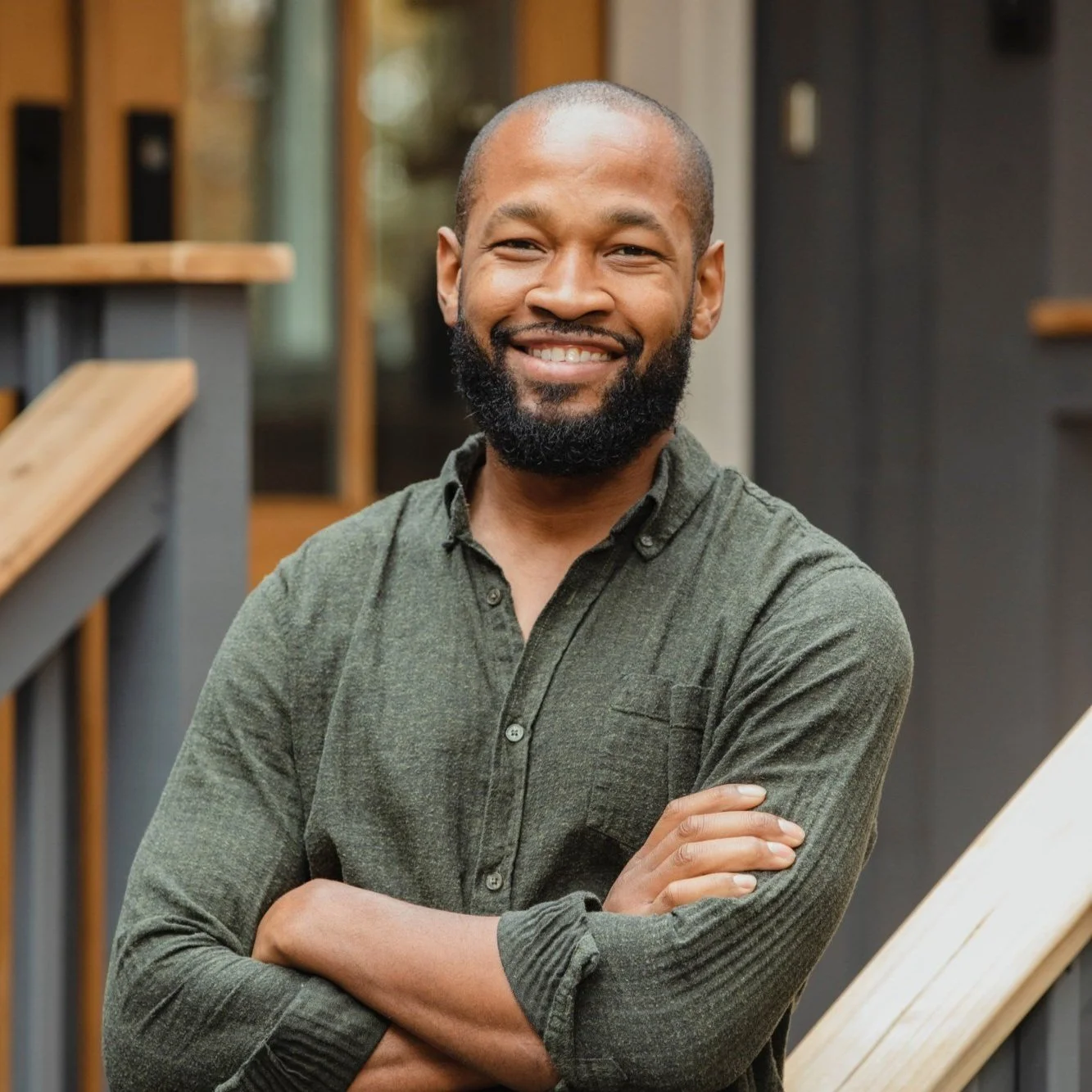 A smiling man with a beard, wearing a green button-up shirt, standing outdoors with arms crossed near a staircase and modern building background.