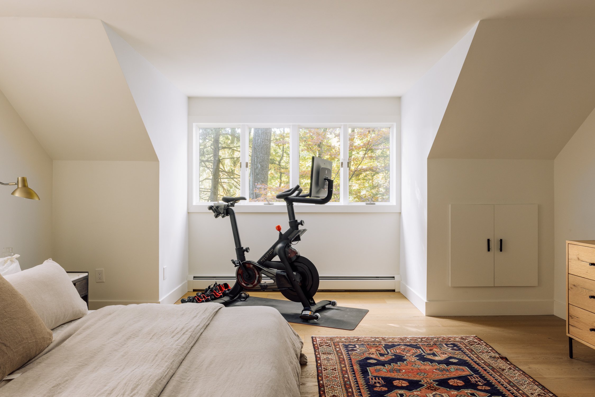 A bedroom with a stationary exercise bike in front of a large window showing trees with autumn leaves.