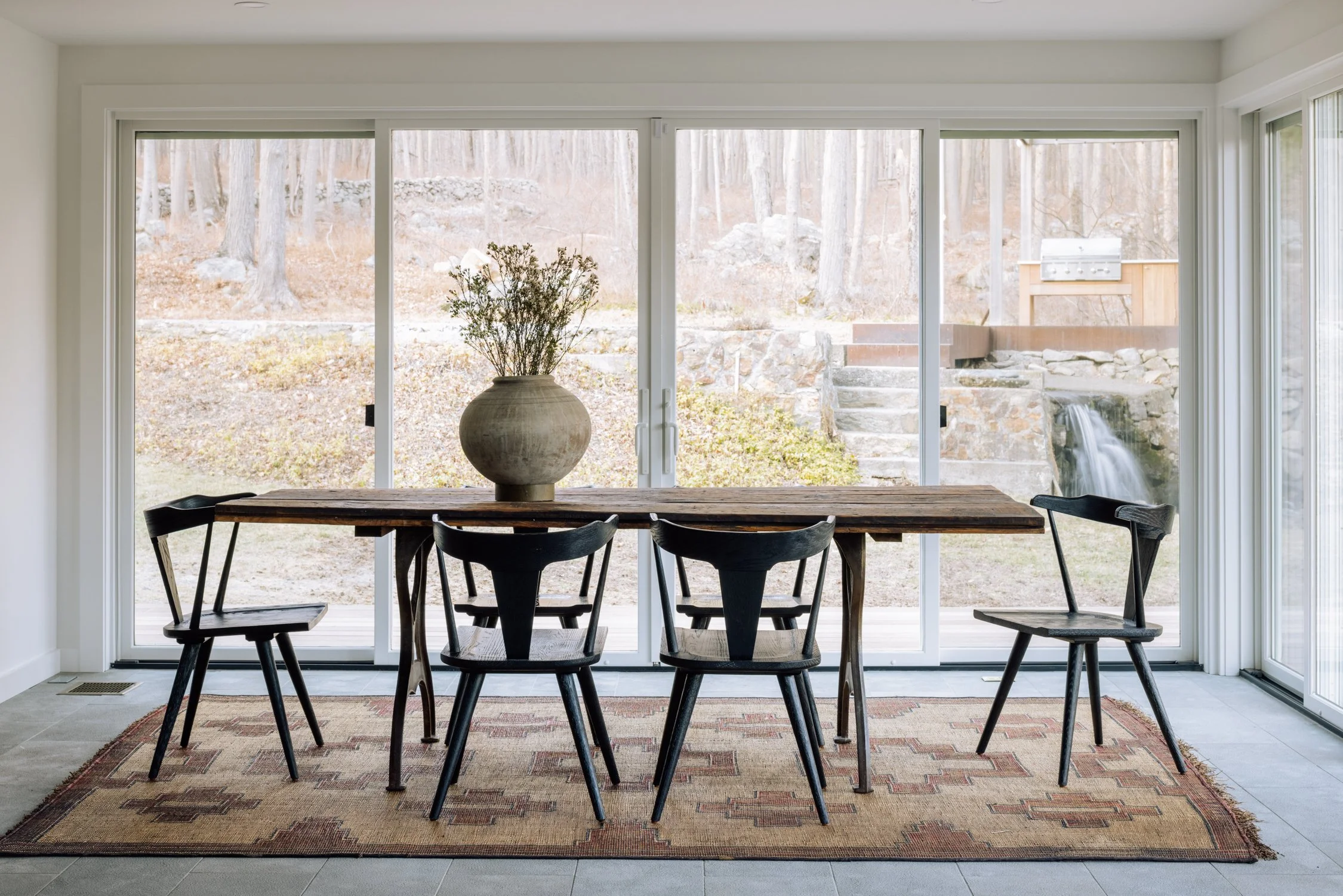 Dining area with a wooden table and six black chairs, a large decorative vase with dried flowers, on a patterned rug, overlooking a backyard with rocks, trees, a waterfall feature, and an outdoor grill.