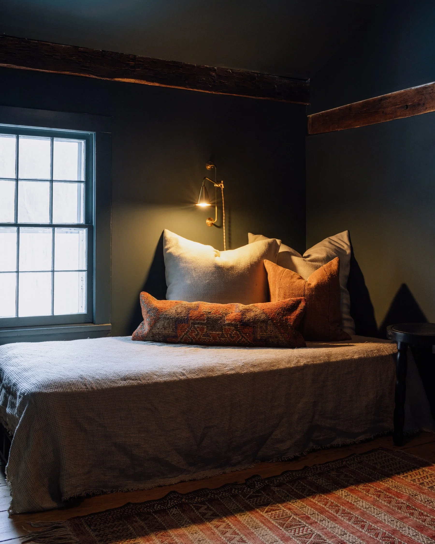 Cozy bedroom with a bed covered in neutral and patterned pillows, a soft beige blanket, a vintage rug, and a window letting in natural light, with a wall-mounted reading light on a dark wall.