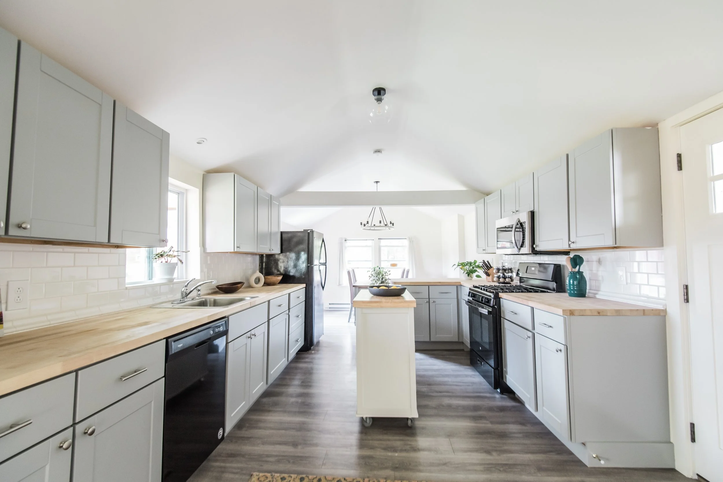 Bright, modern kitchen with light gray cabinets, wooden countertops, white subway tile backsplash, black appliances, and a small central island with a wooden top. There is a window above the sink and a dining area with large windows at the far end.