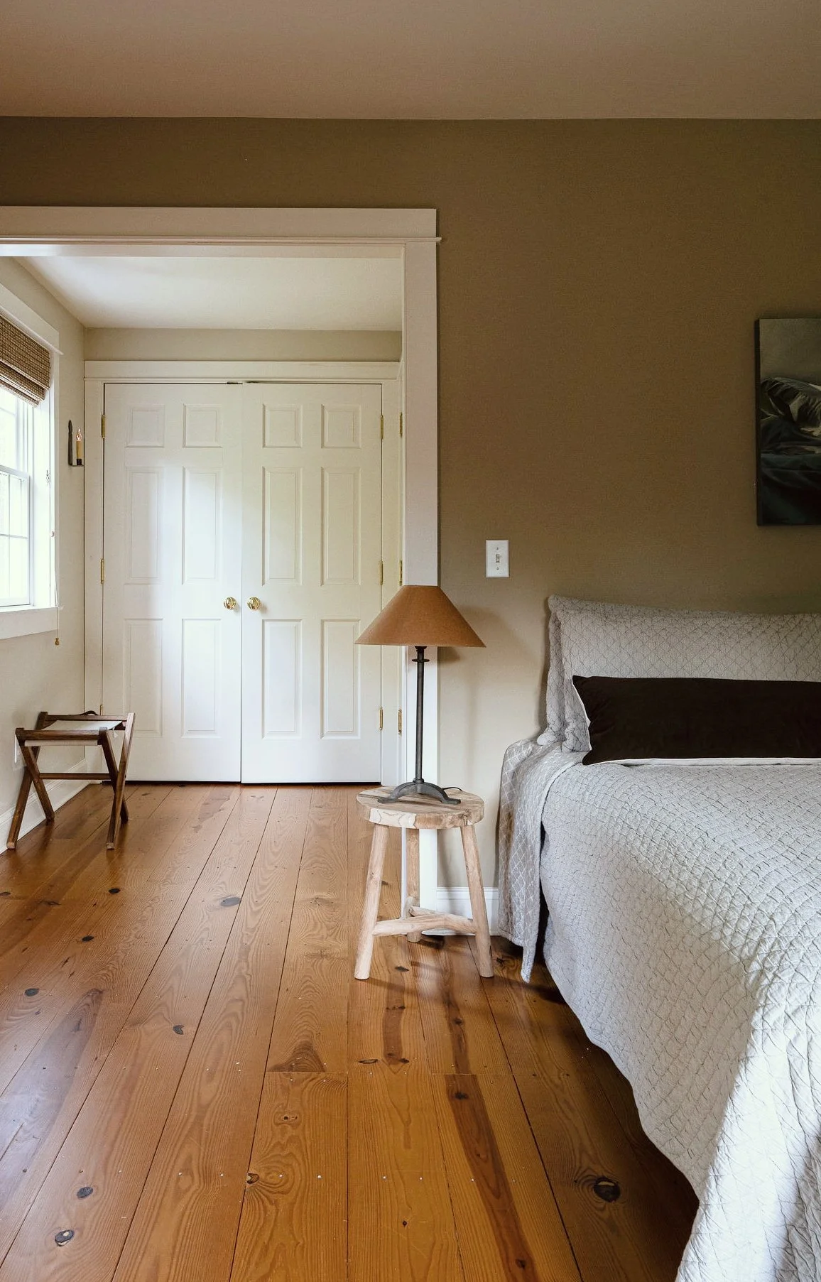 Bedroom with wooden floor, beige wall, white double closet doors, window with blinds, bedside lamp on a small wooden stool, and part of a bed with dark pillow and beige quilted bedding in the corner.