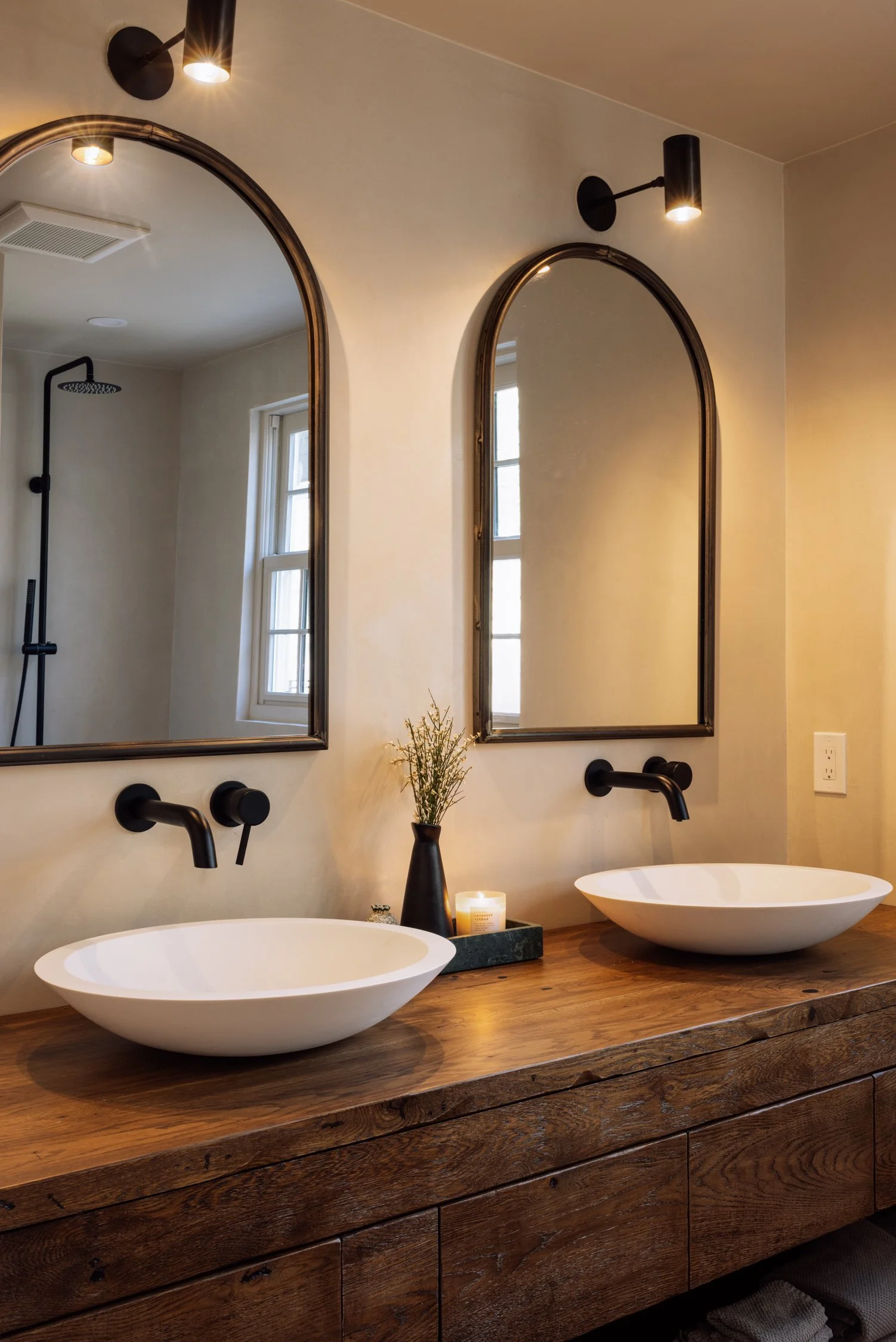 Bathroom double vanity with wooden countertop, oval ceramic sinks, black wall-mounted faucets, two arched mirrors, black wall sconces, a small black vase with dried flowers, and a lit candle.