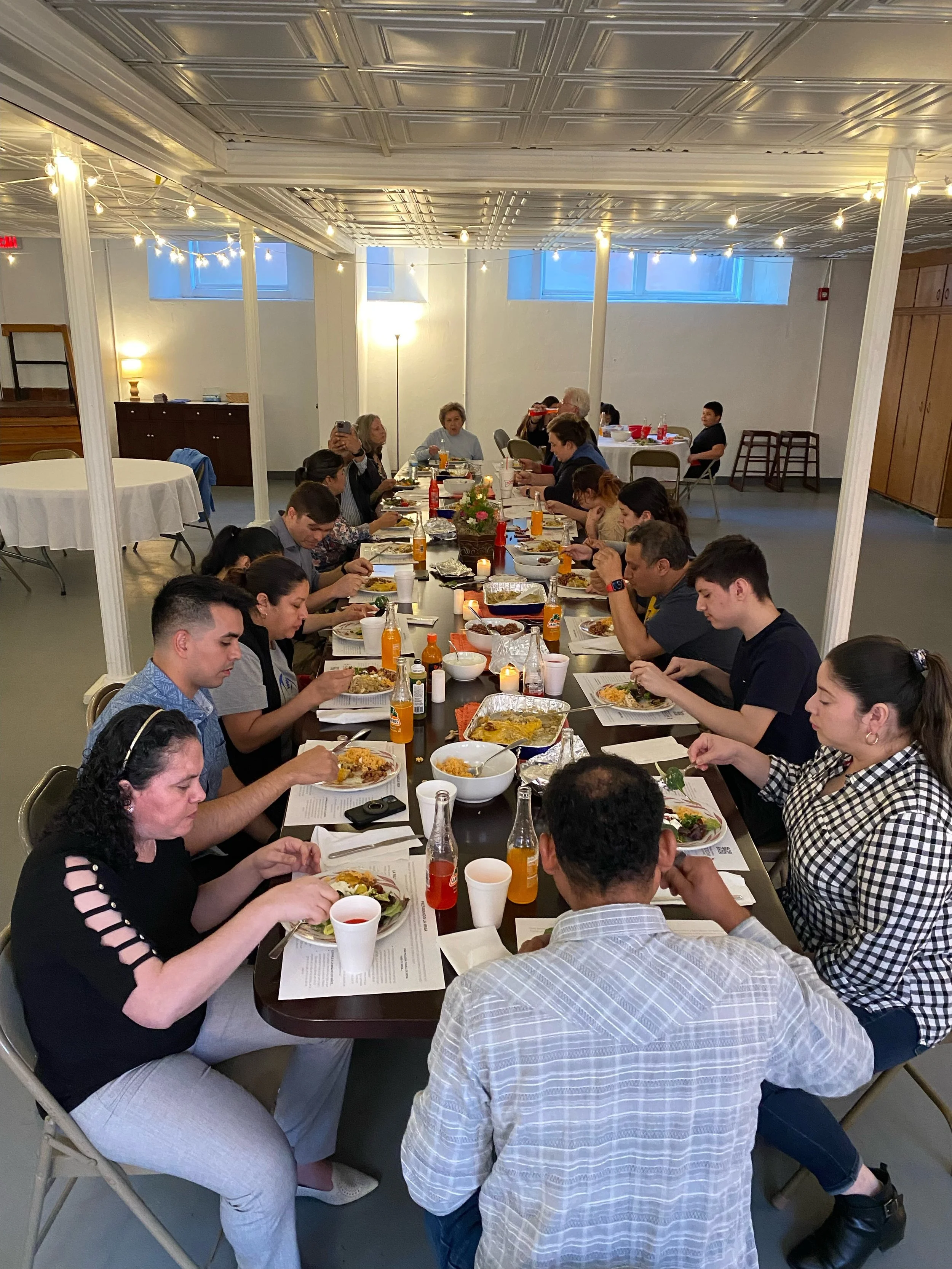 A group of people gathered around a long table enjoying a meal in a large, well-lit room with string lights hanging from the ceiling. People are eating, drinking, and engaging in conversation.