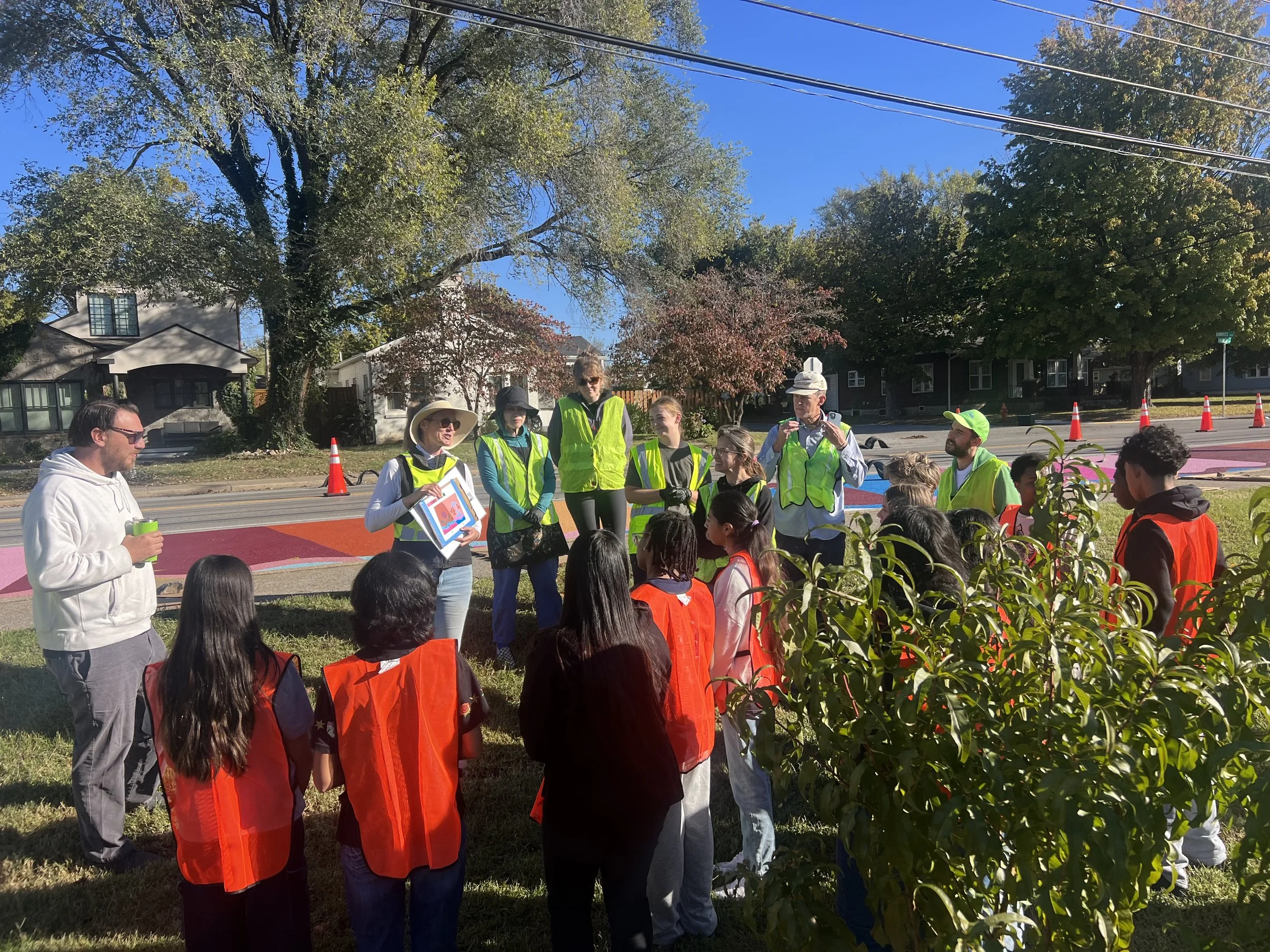 A diverse group of children and adults gathered outdoors around a woman showing a colorful picture, with some wearing bright orange safety vests and others wearing hats. There are trees and houses in the background on a sunny day.