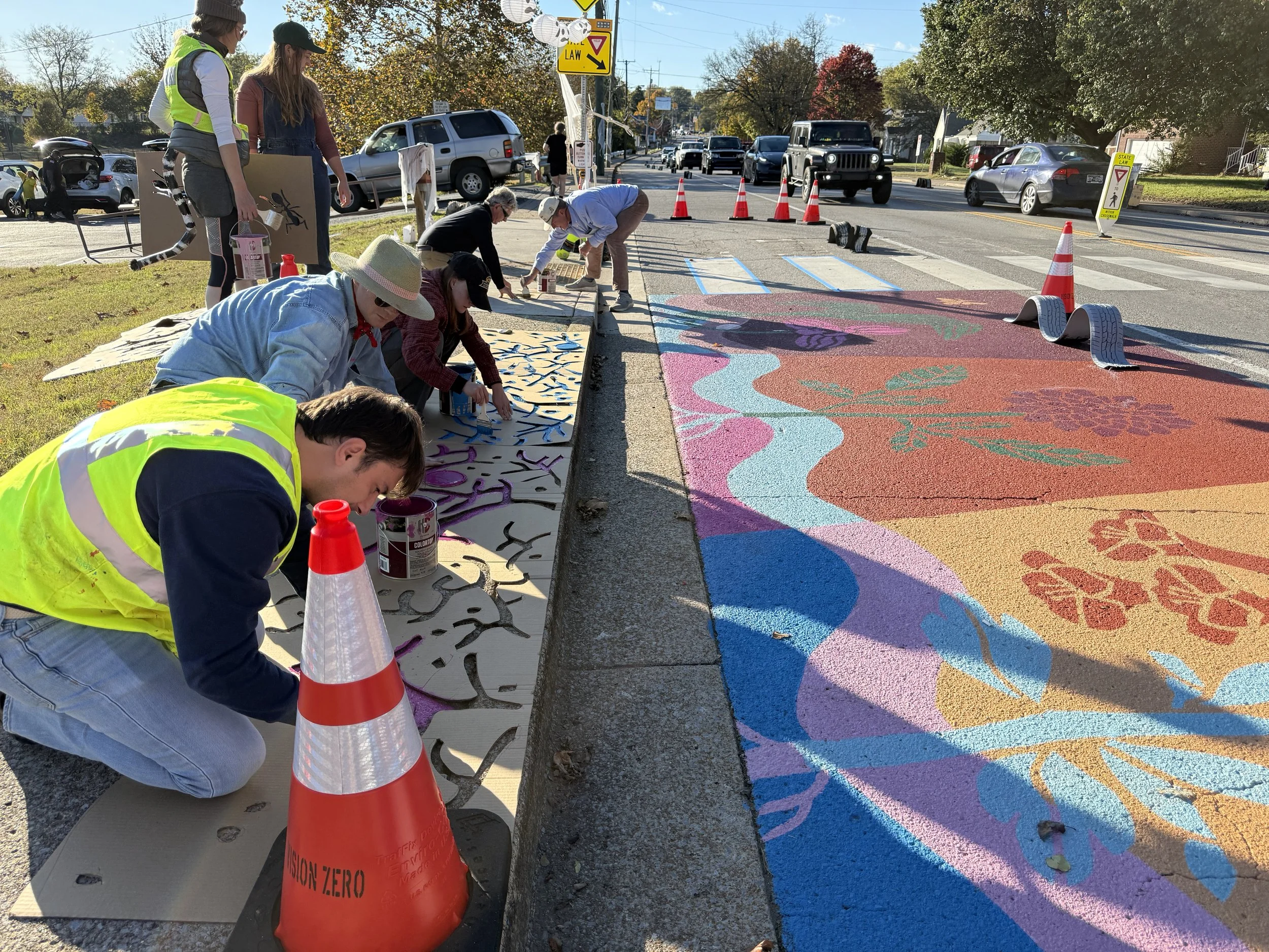 Several people are painting a colorful mural on the sidewalk next to a busy street with cars passing by. Traffic cones and signs are placed around the work area, and the sky is clear.
