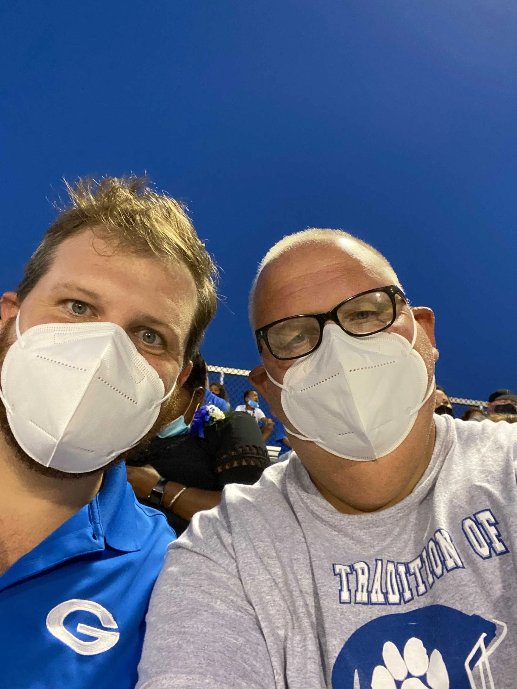 Dr. Kuzma Jr. and Dr. Kuzma Sr. wearing face masks and casual shirts taking a selfie outdoors during evening. One man has light brown hair and a blue shirt with a logo, the other is bald, glasses, and a gray shirt with a blue logo.