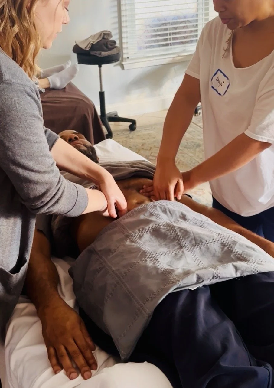 Two women are giving an abdominal massage treatment to a man; one woman is coaching the other as they are in a continuing education class for massage therapists at the NC Massage School