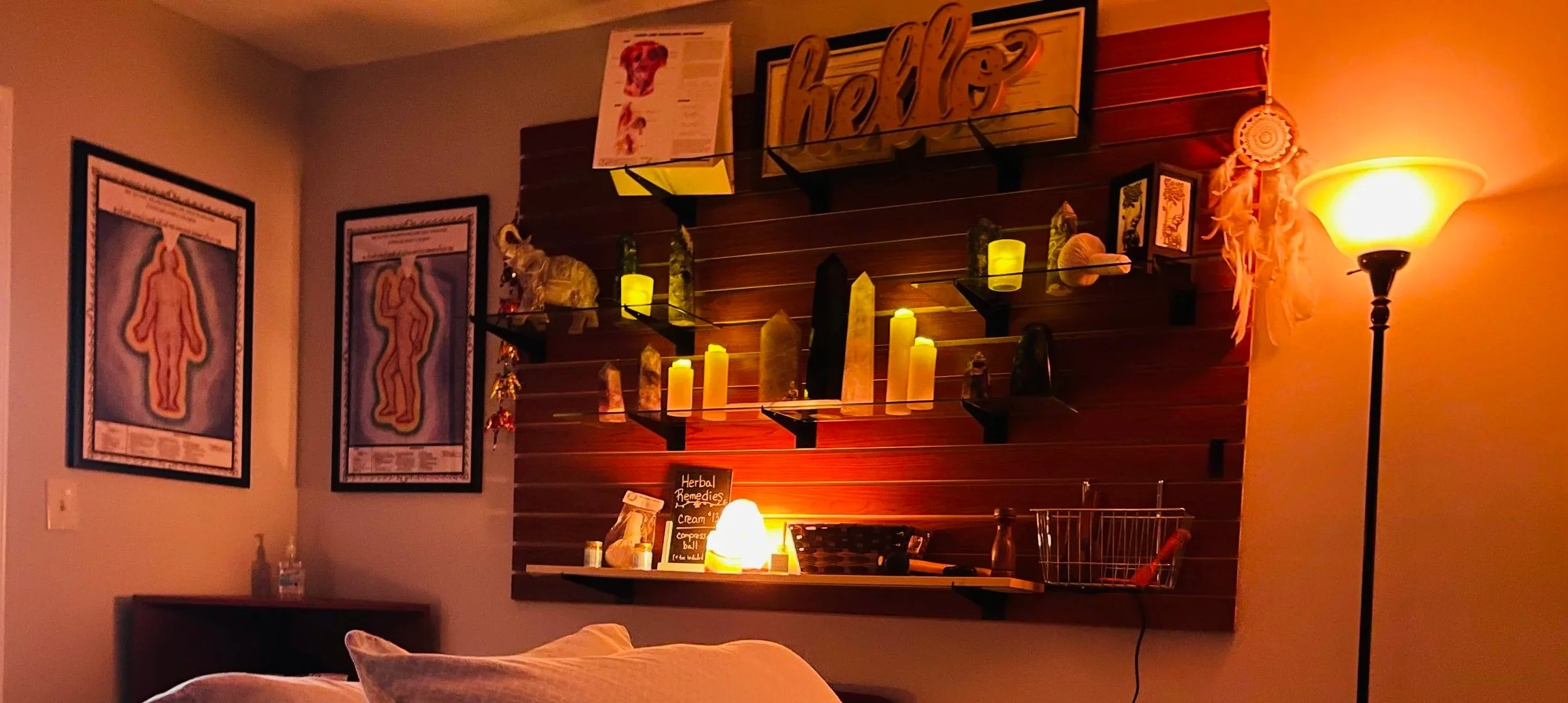 A cozy massage therapy room with two framed anatomical posters on the wall, a wooden shelf decorated with candles, crystals, and a small plant, and a warm yellow floor lamp. The shelf includes a small chalkboard menu offering herbal remedies.