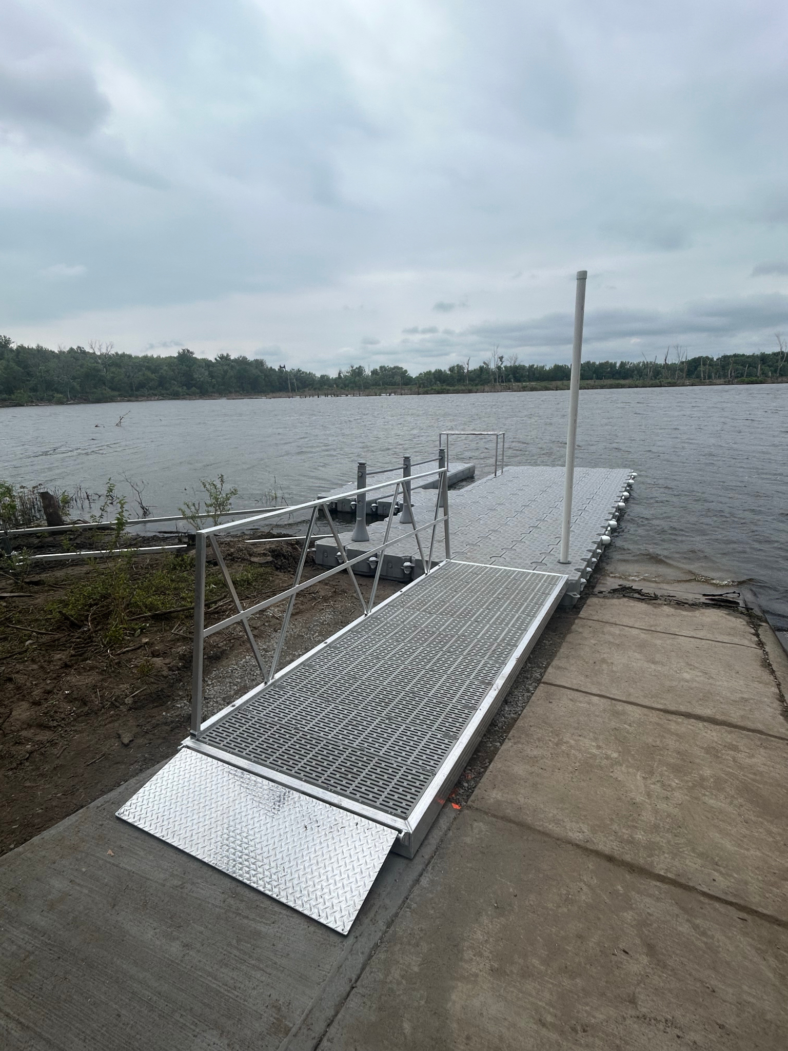 Metal dock ramp extending into a lake on a cloudy day, surrounded by trees in the distance.