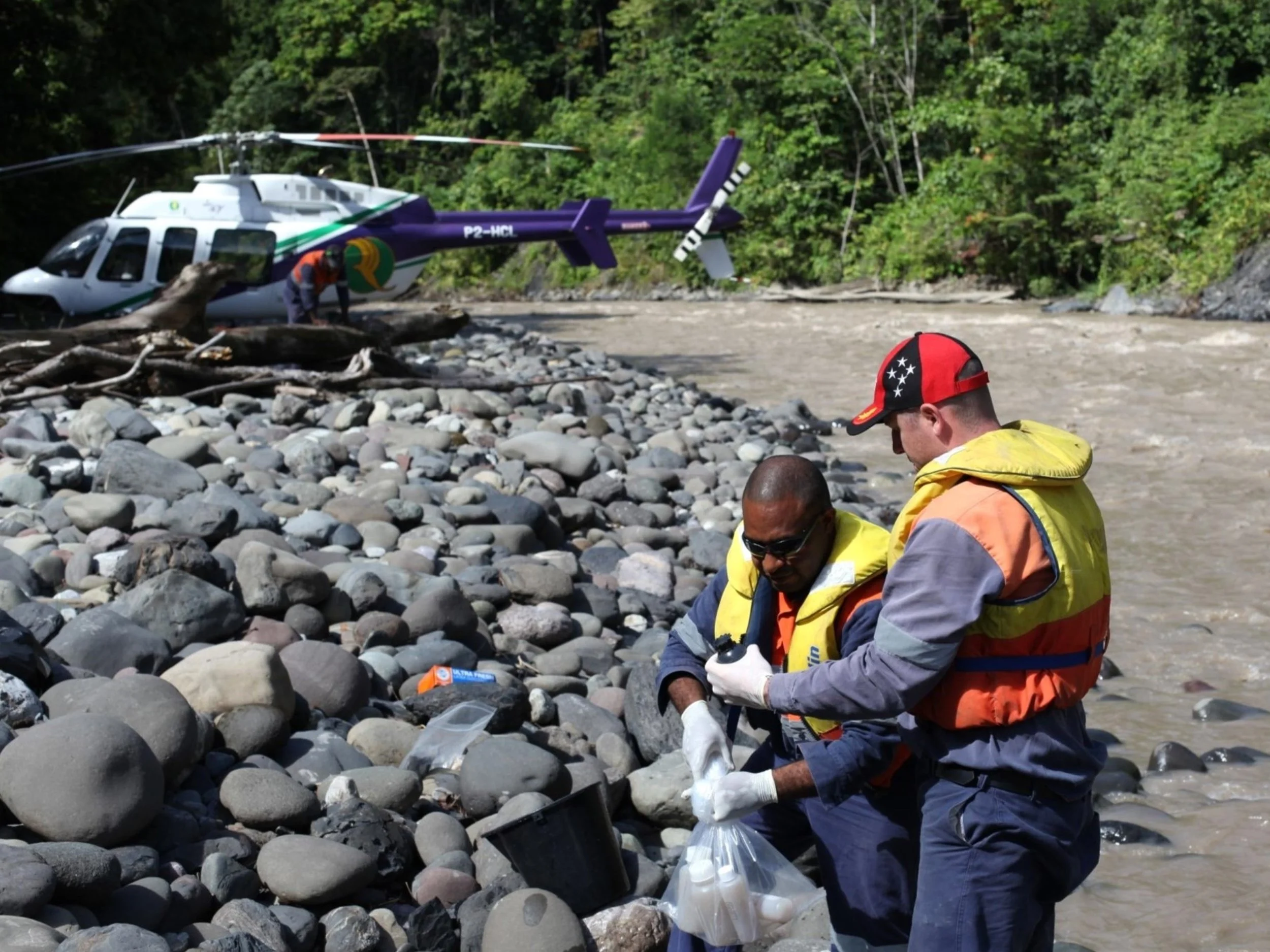 Rescue workers examining evidence on a rocky riverbank with a helicopter in the background.