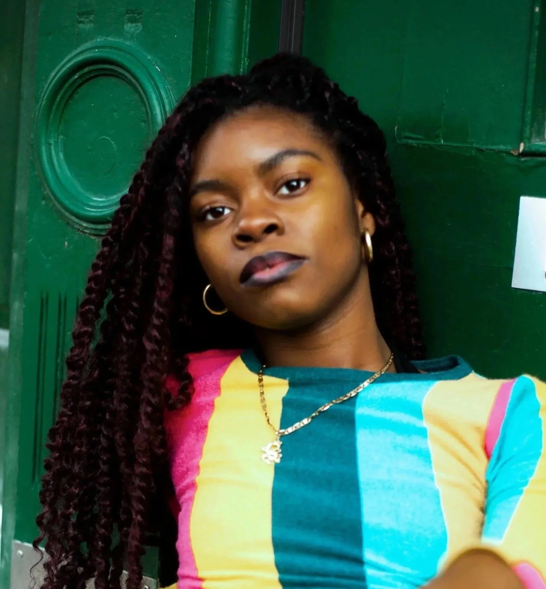 Jeune femme noire avec des cheveux torsadés portant un t-shirt coloré, elle pose devant une porte verte.