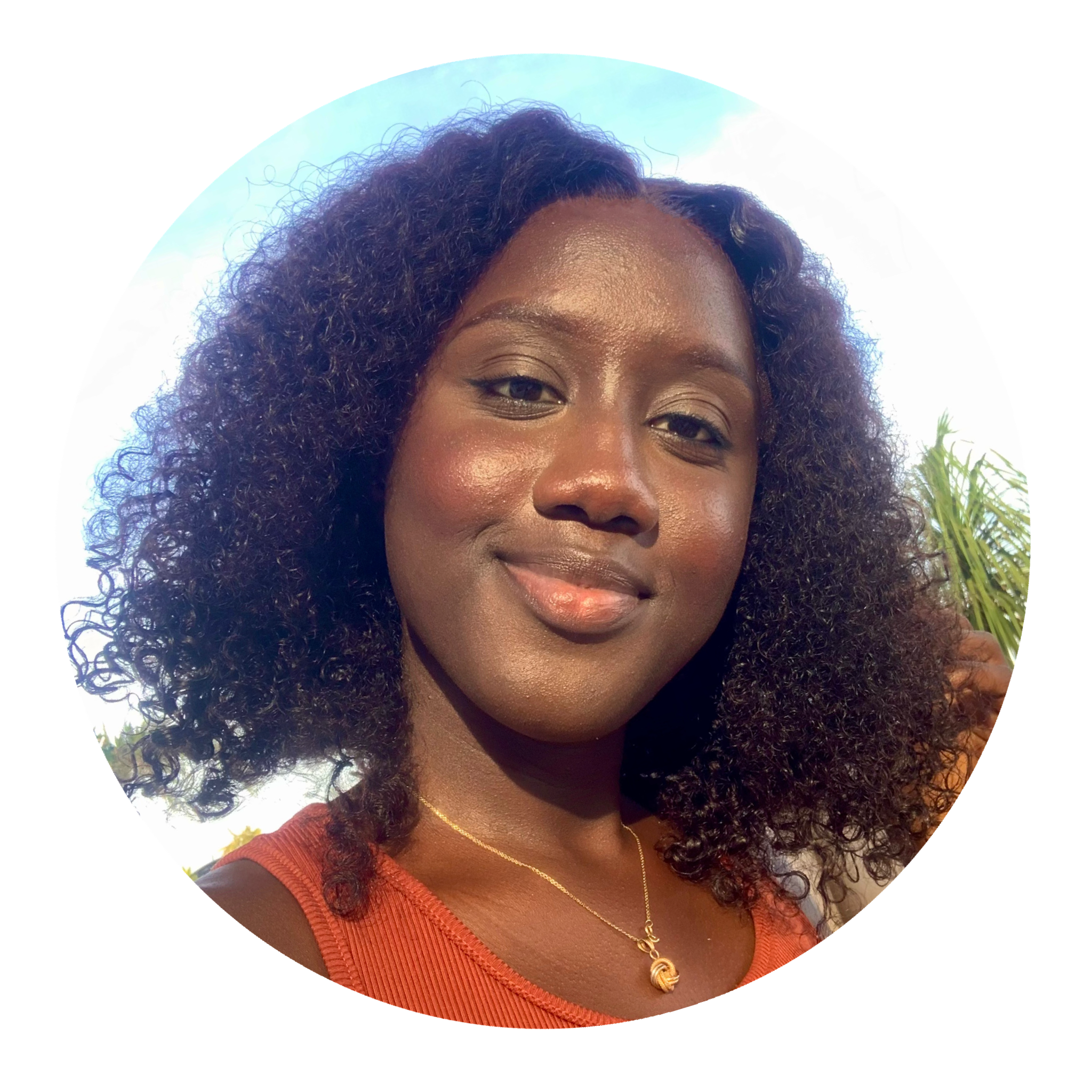 Portrait d'une femme noire souriante avec des cheveux bouclés, portant un haut orange et un collier doré, avec un ciel bleu et des plantes en arrière-plan.