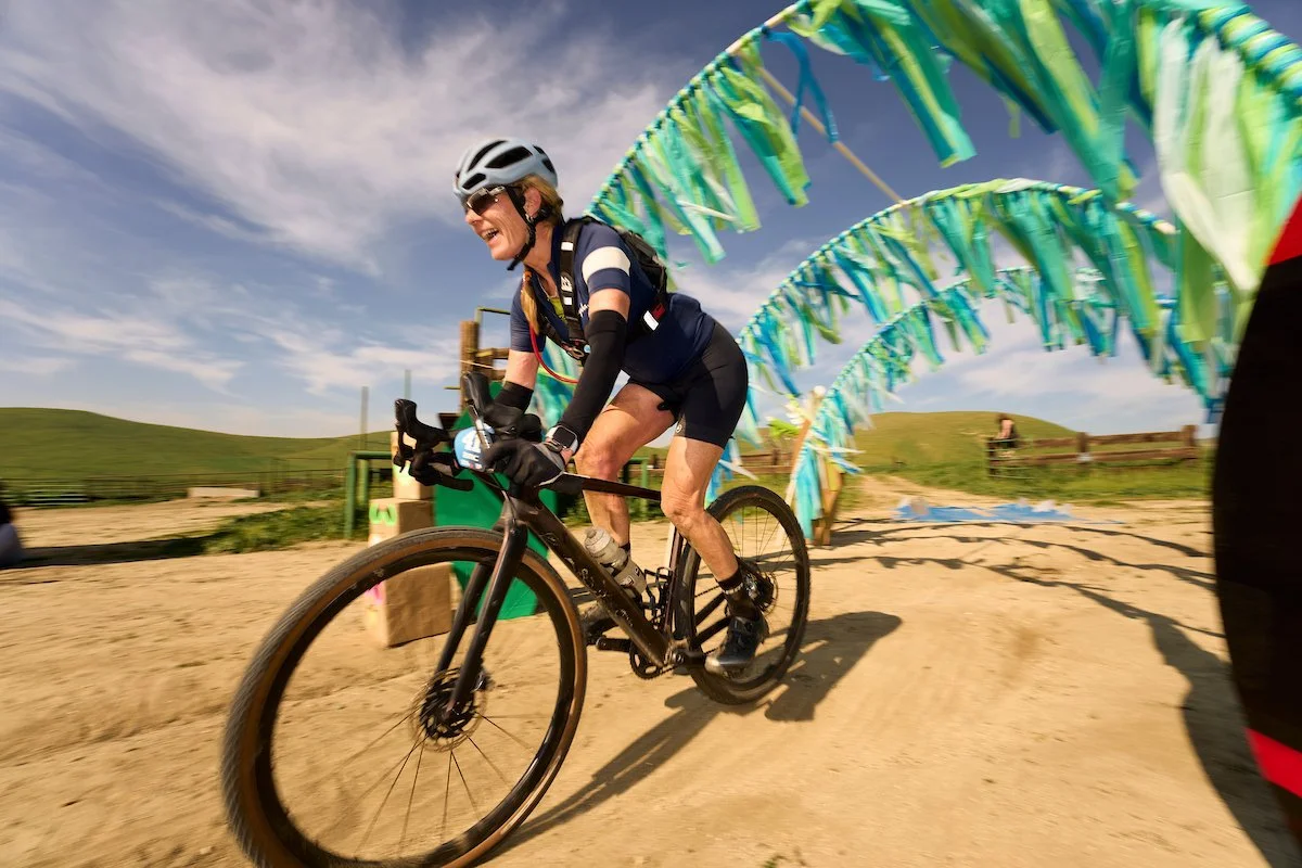 Two cyclists riding on a dirt road between rows of green bushes, with mountain range in the background, one of the cyclists raising both hands in celebration.