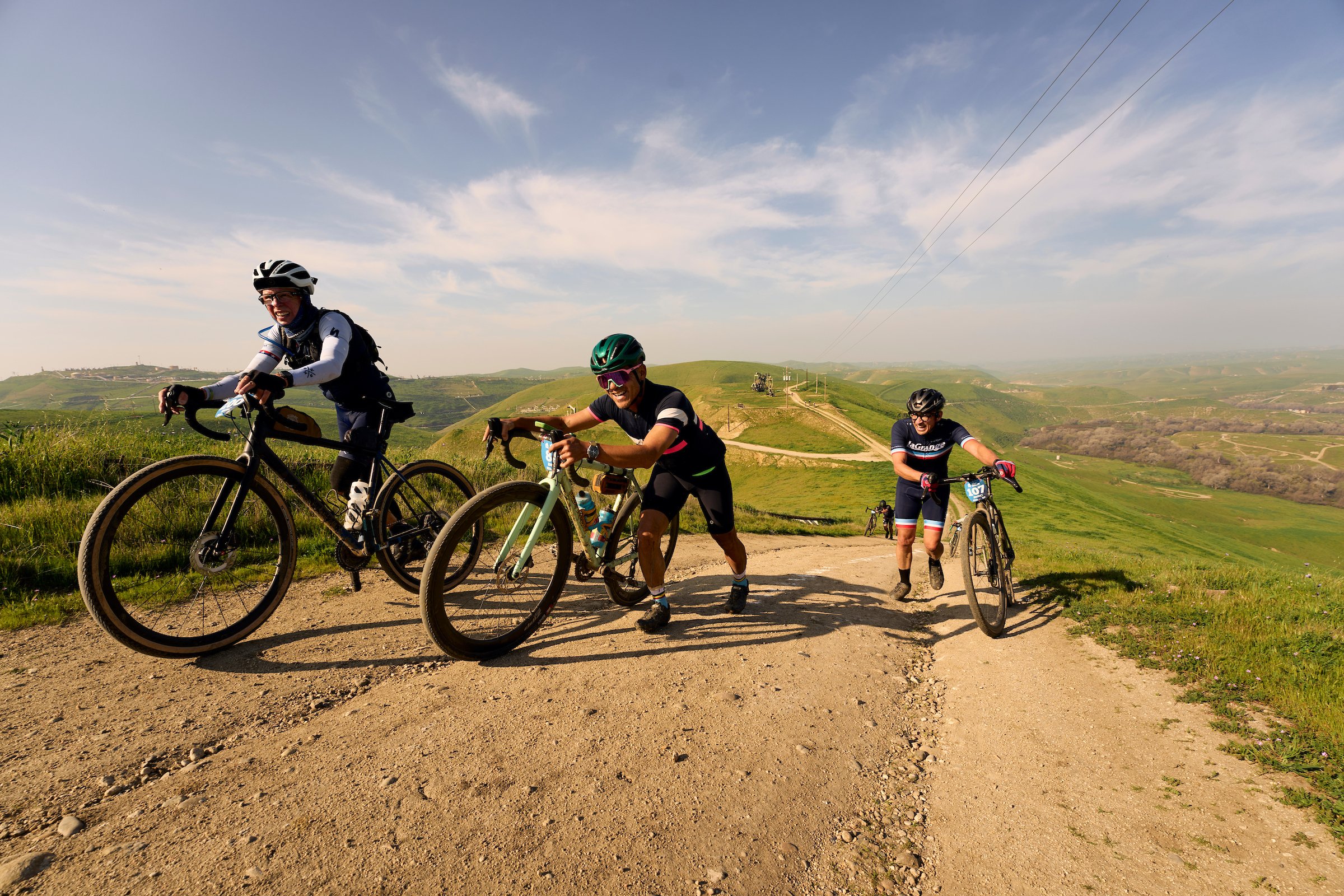 Four cyclists riding gravel bikes through a grassy field with rolling hills and mountains in the background under cloudy sky.