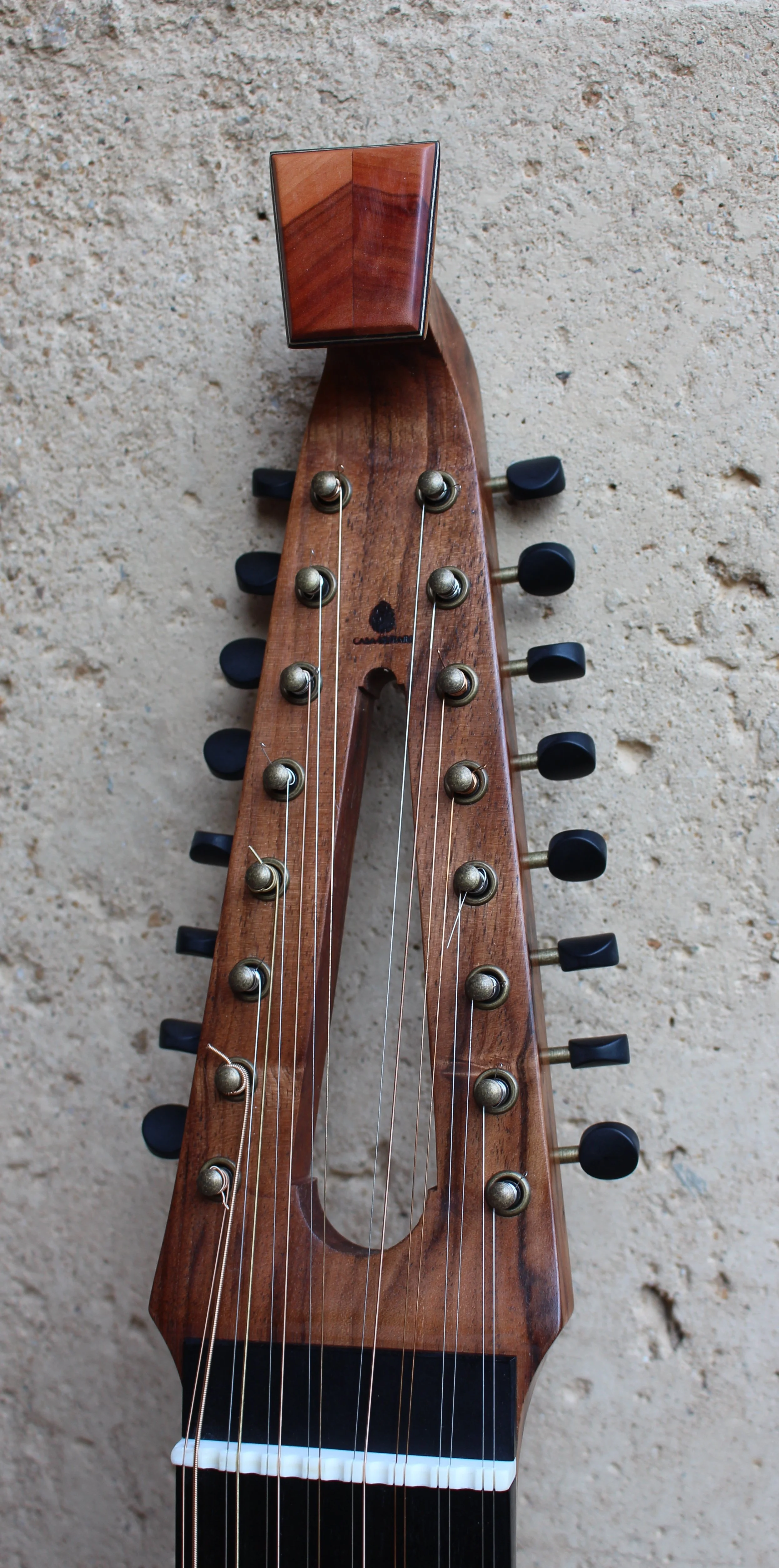 Tête d'un instrument de musique en bois avec des cordes, probablement un oud, sur un mur en pierre.