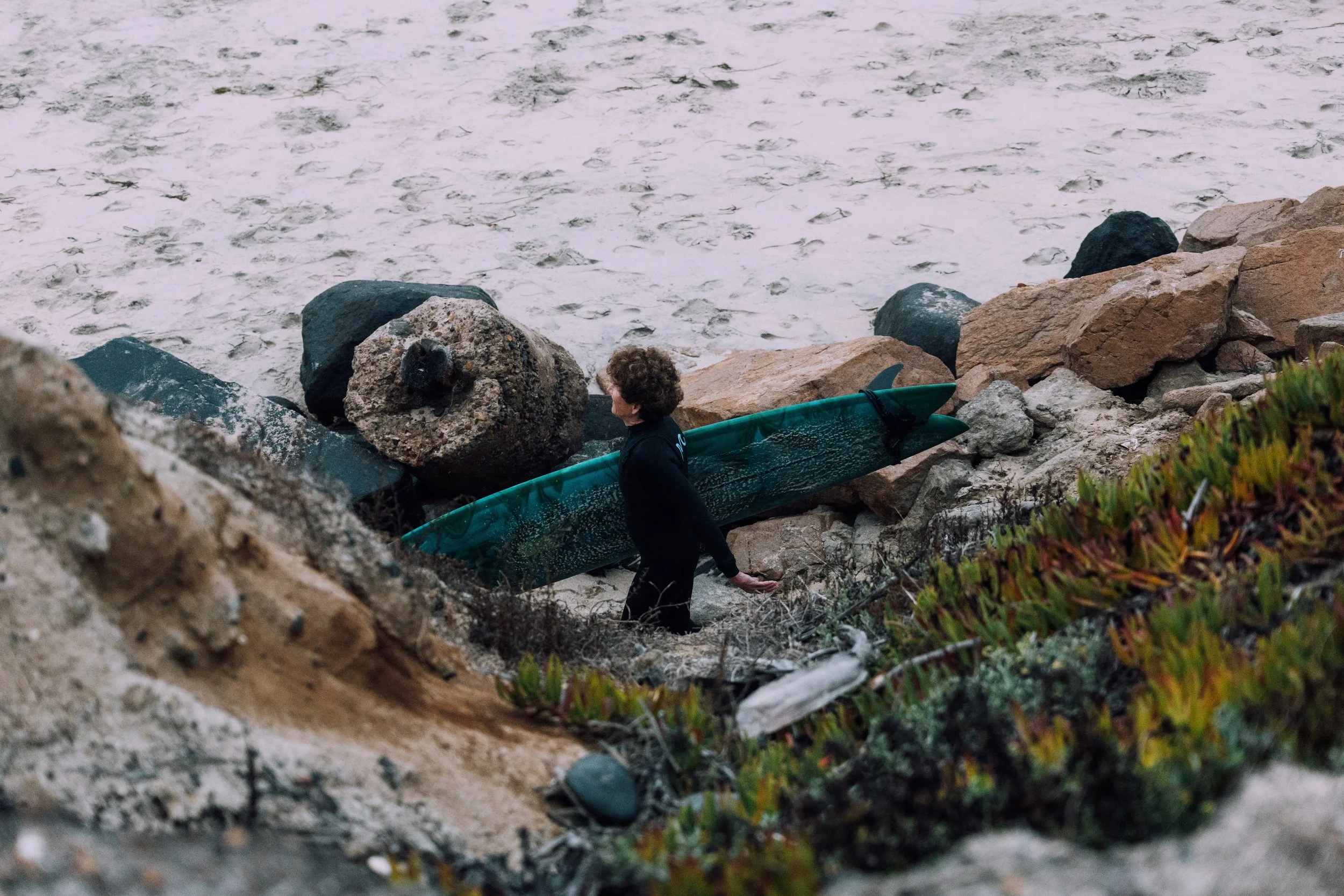 A person with curly hair carrying a green surfboard and walking on a rocky beach with sand and vegetation.