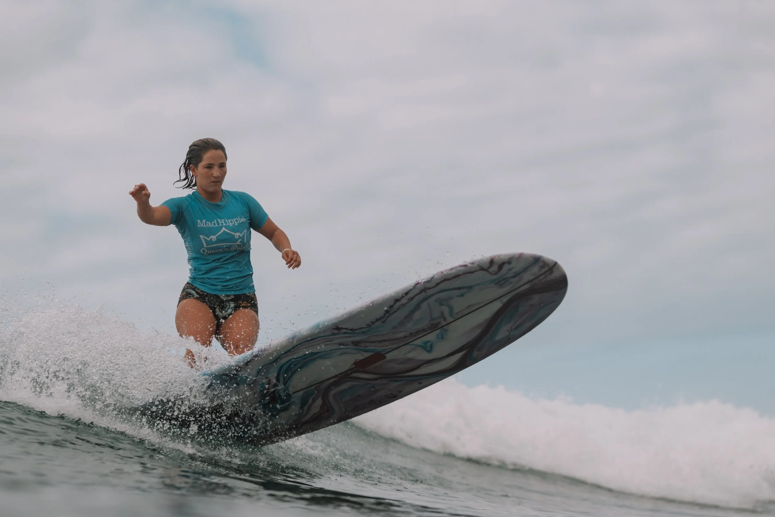 Woman surfing on a wave, wearing a blue t-shirt with the text 'Mad Hippie Queen of' and black shorts, overcast sky in background.