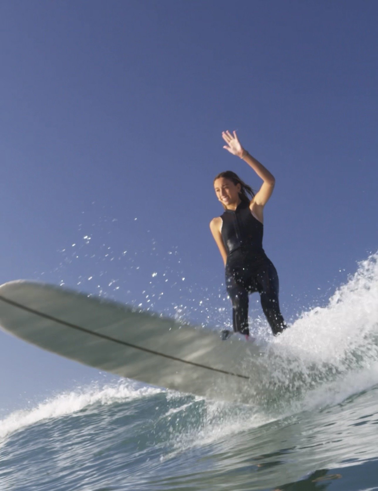 A woman surfing on a wave in the ocean, waving and smiling, wearing a black wetsuit, under a blue sky.
