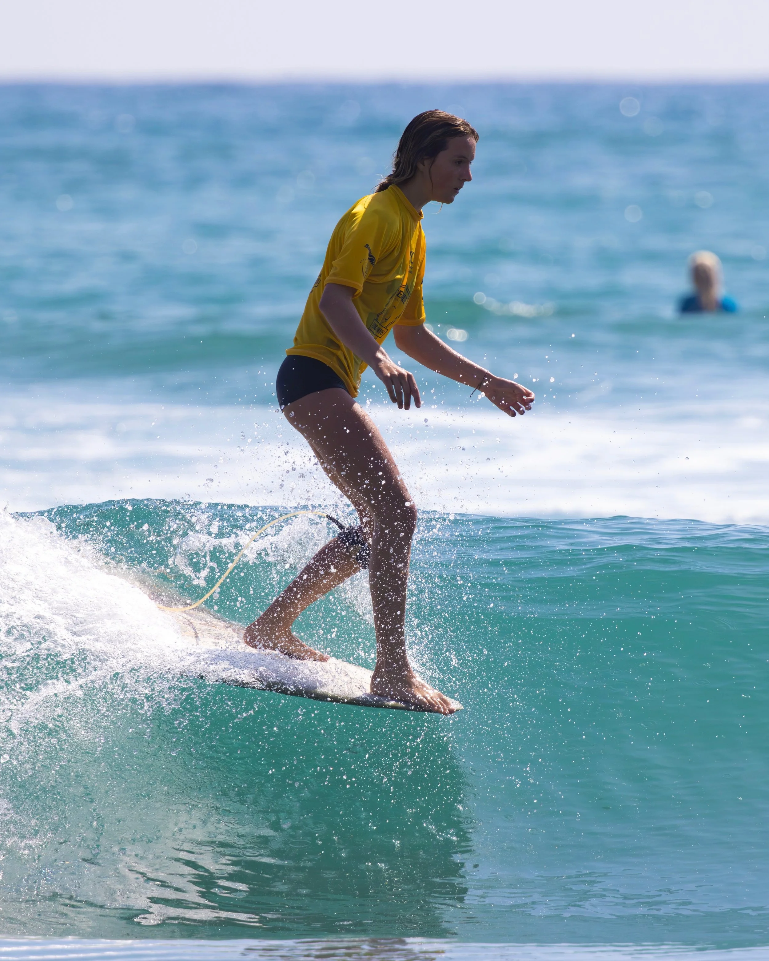 A person in a yellow shirt surfing on a wave in the ocean with another swimmer in the background.