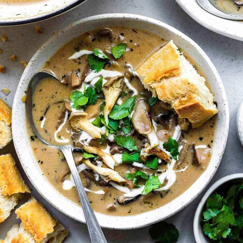 A Creamy soup base with large oyster mushroom portions sprinkled on top served alongside a perfectly crusty baugette slice.
