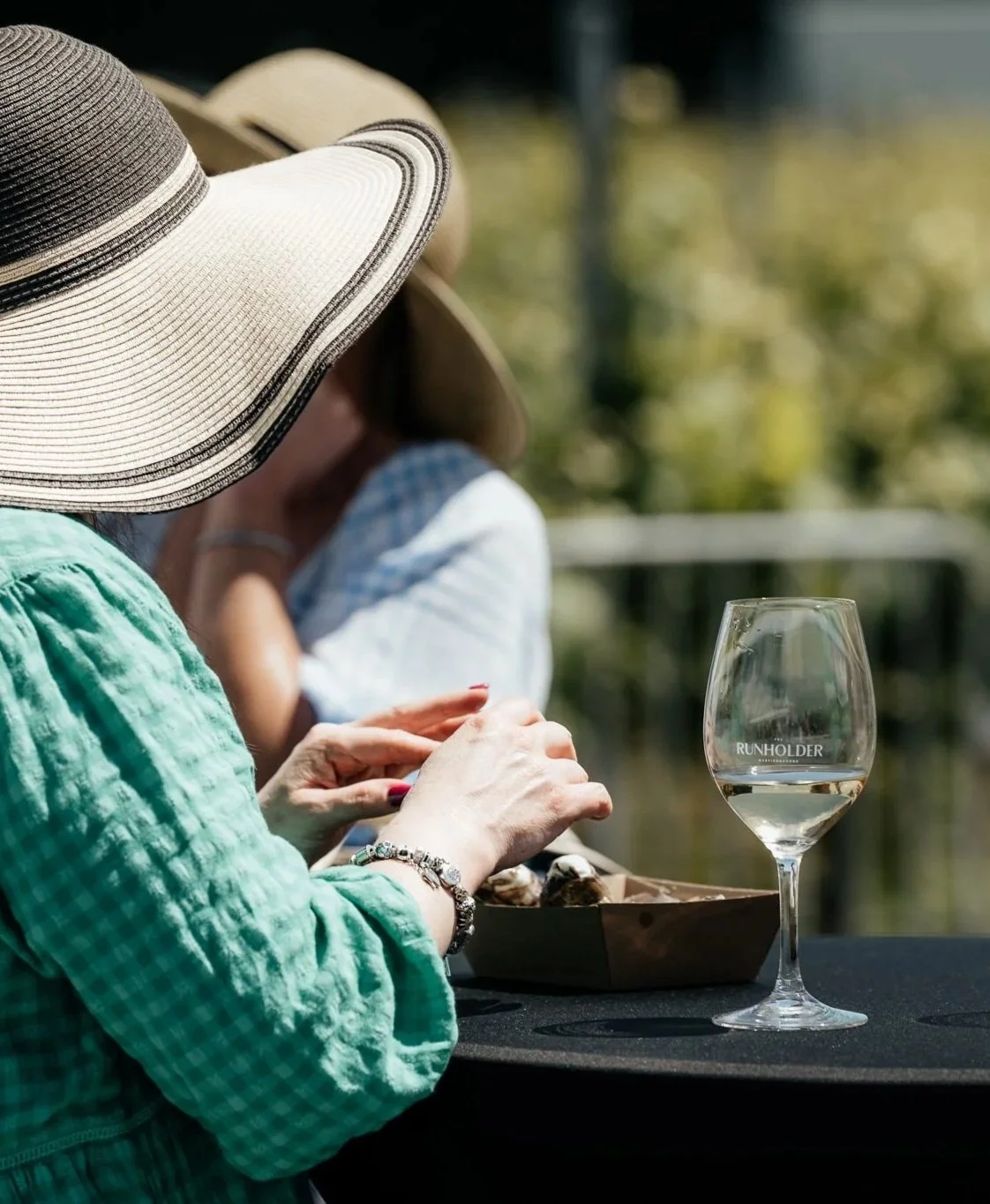 Person wearing a large sun hat and a green blouse sitting at a table outdoors, with a glass of white wine and a basket of food in front of them. Toast Martinborough at The Runholder.