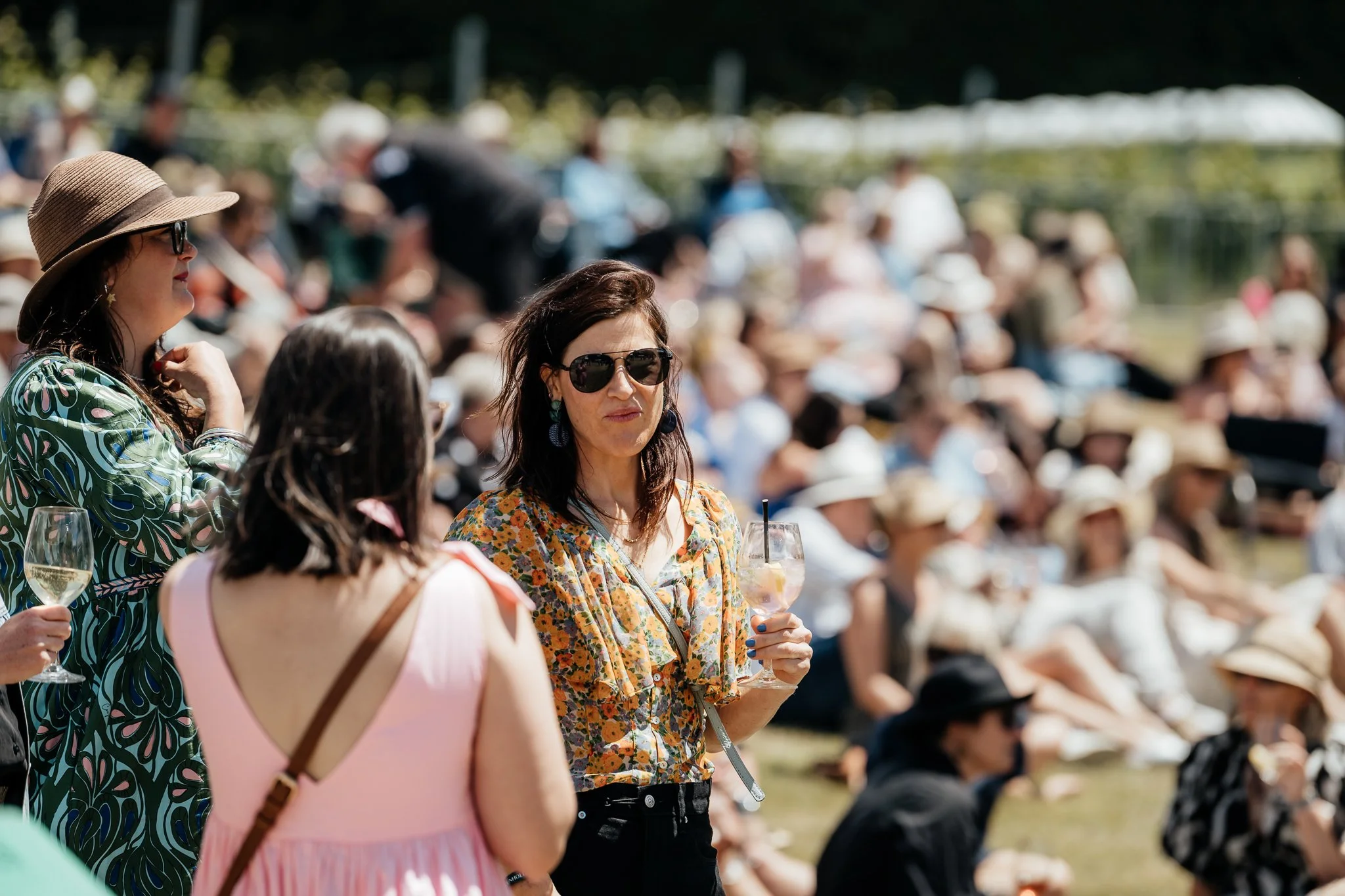Group of people attending an outdoor event, Toast Martinborough, sitting on grass and wearing summer clothing and hats, some holding drinks, with a woman in the foreground wearing sunglasses and a floral top, holding a drink.
