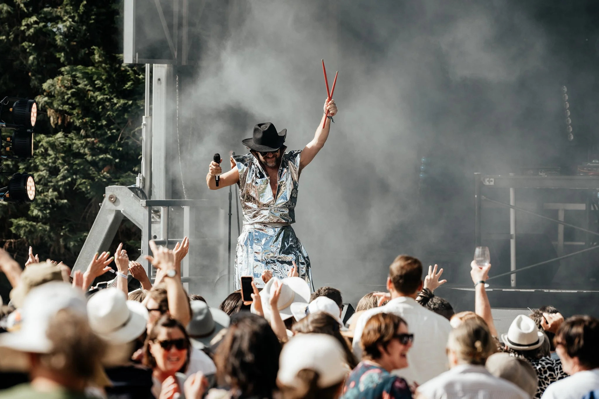 Sweet Mix Kids at The Runholder. A performer on stage at an outdoor event, wearing a shiny silver outfit and black hat, holding a microphone in one hand and drumsticks in the other, with a crowd of people in sun hats and sunglasses cheering.