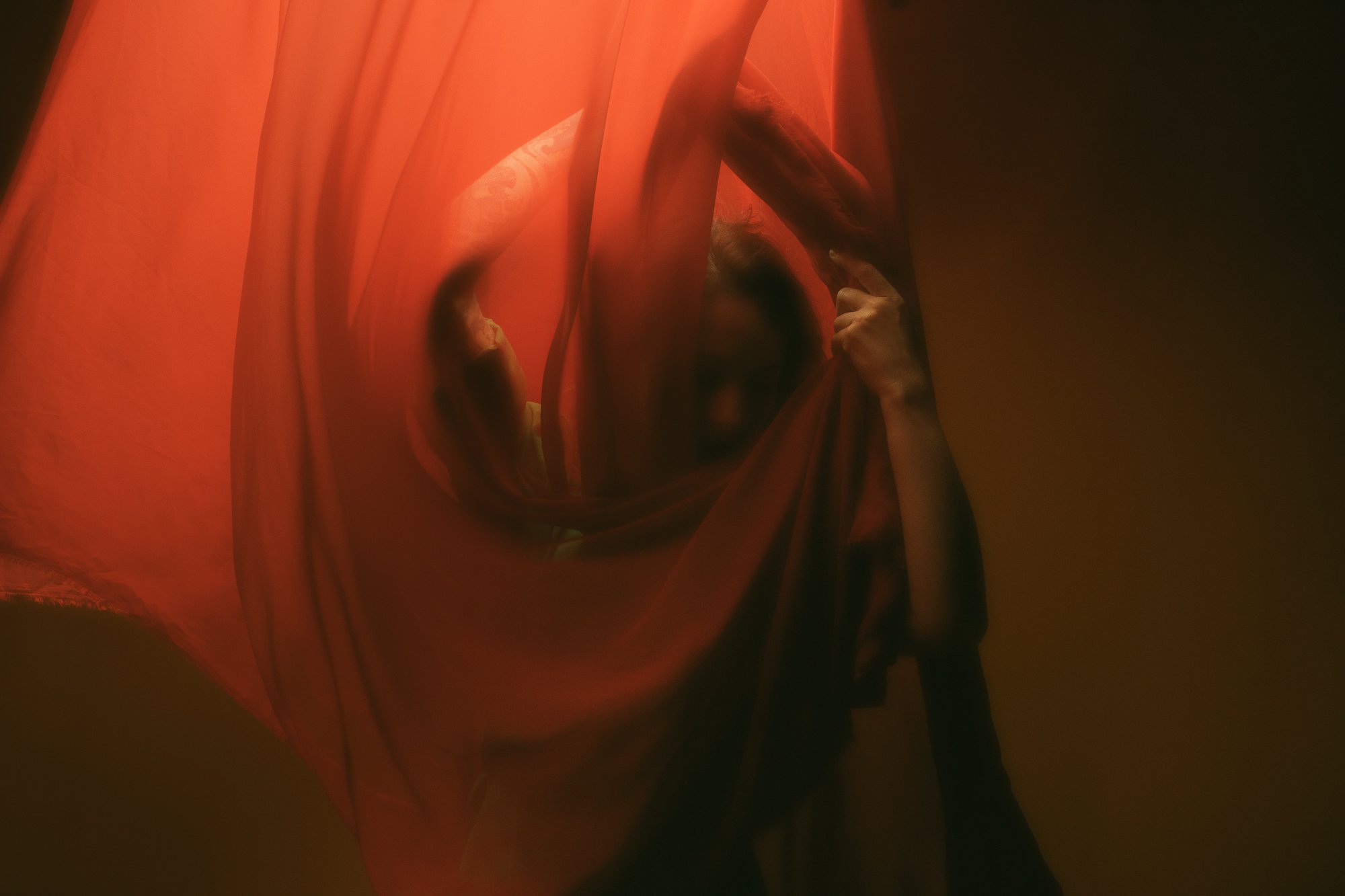 A person partially hidden behind a red curtain, holding it with one hand and peering out from behind it.