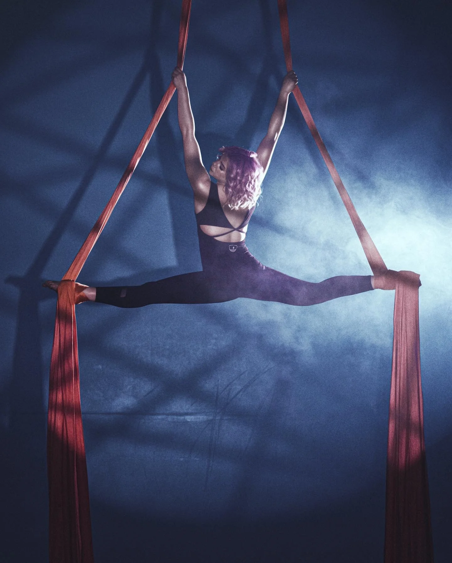 A woman with pinkish-purple hair performing an aerial split on Silks in a silks class at Academy of Acrobatic Sports in Richland